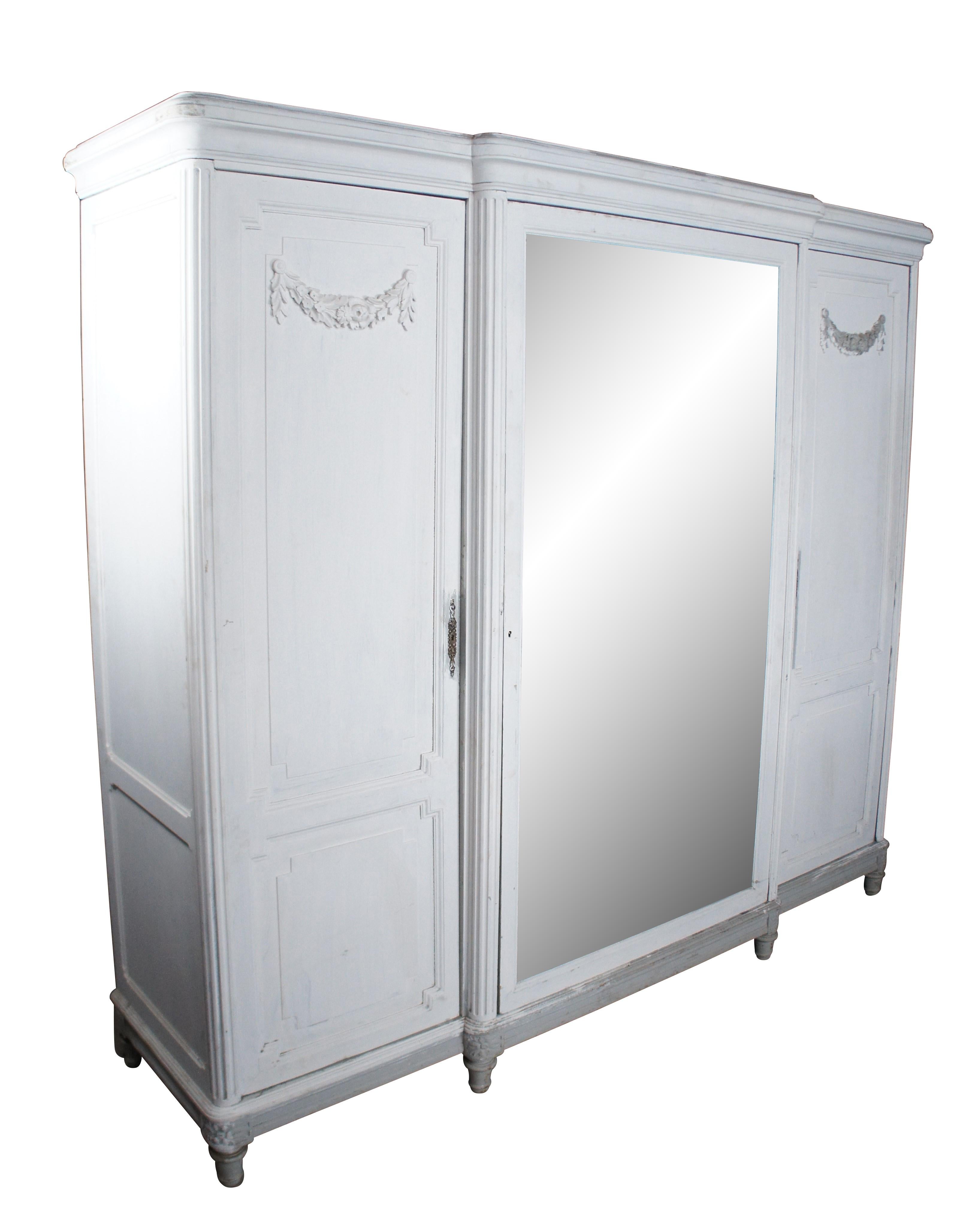 Antique French Provincial knockdown armoire / wardrobe / closet.  Made of oak featuring a chic white painted finish with large central beveled vanity / dressing mirror flanked by two cabinets with Neoclassical carved accents.

Dimensions:
89