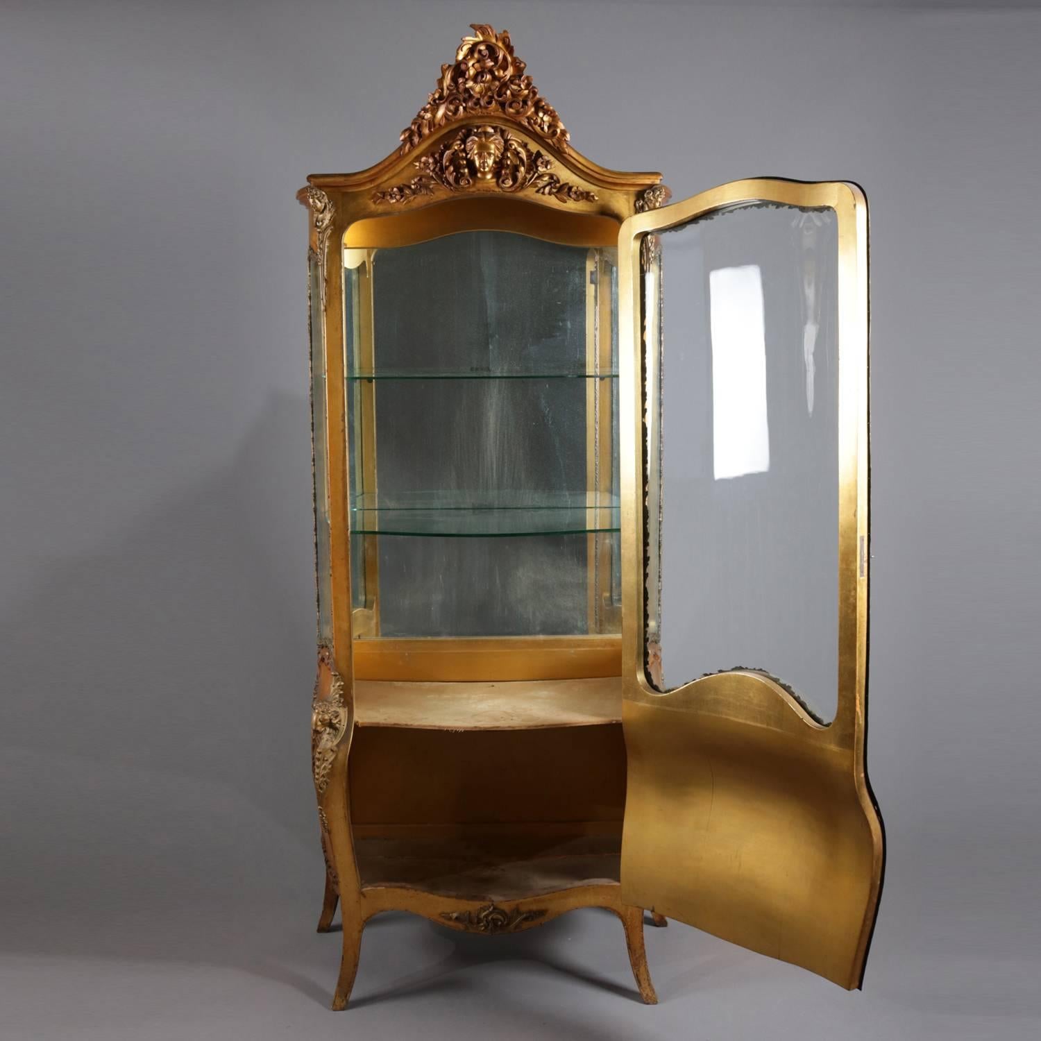 Antique French Rococo figural vitrine features carved giltwood construction with scroll and foliate cartouche above Jenny Lind mask crest, bow front single door with bombe form lower and central Jenny Lind mask opens to reveal mirror back display