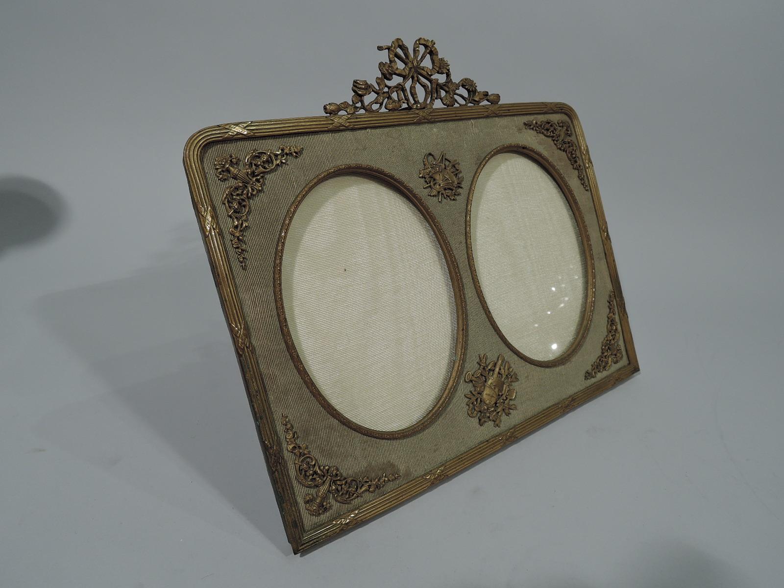 French Rococo Revival gilt-bronze double picture frame. Rectangular with curved corners, reeded rim, and bow-tied floral crown. Two vertical oval windows and ribbed textile ground applied with 18th-century-style garden and music trophies. Subdued,