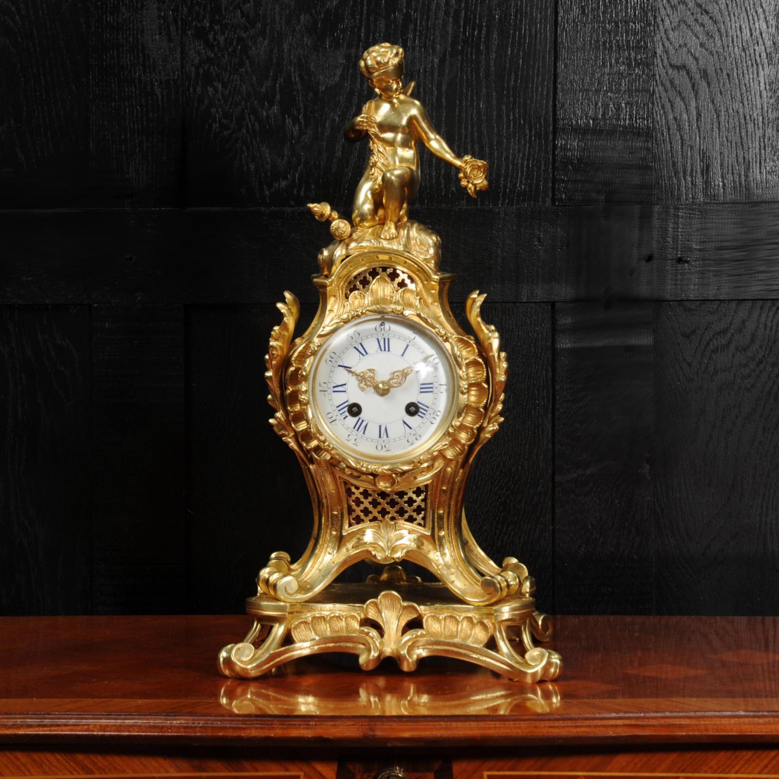 A stunning original antique French Rococo clock. The case made of finely gilded bronze in the Louis XV style of bold scrolling acanthus forming a waisted case, standing upon a base. Fabric fretted panels lighten the design. Cupid is depicted as a