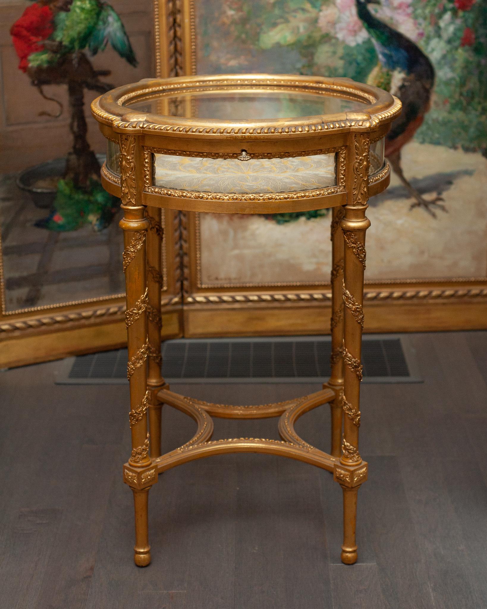 A French antique round gilded display table with ornately carved legs. The interior is lined with a silk damask. The top lifts up and can be locked with a key.