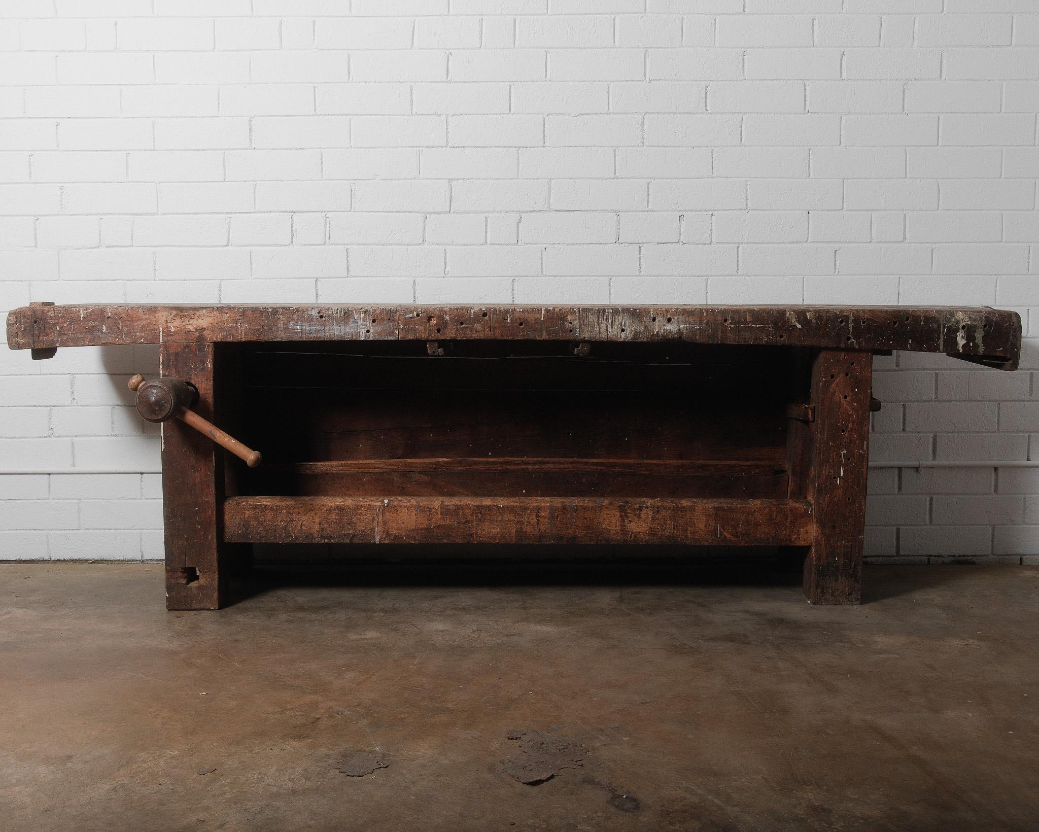 This 19th-century French carpenter’s workbench is a true relic of craftsmanship, built for function and endurance. The thick, well-worn tabletop bears the marks of countless woodworking projects, featuring original tool holes and a rich patina from