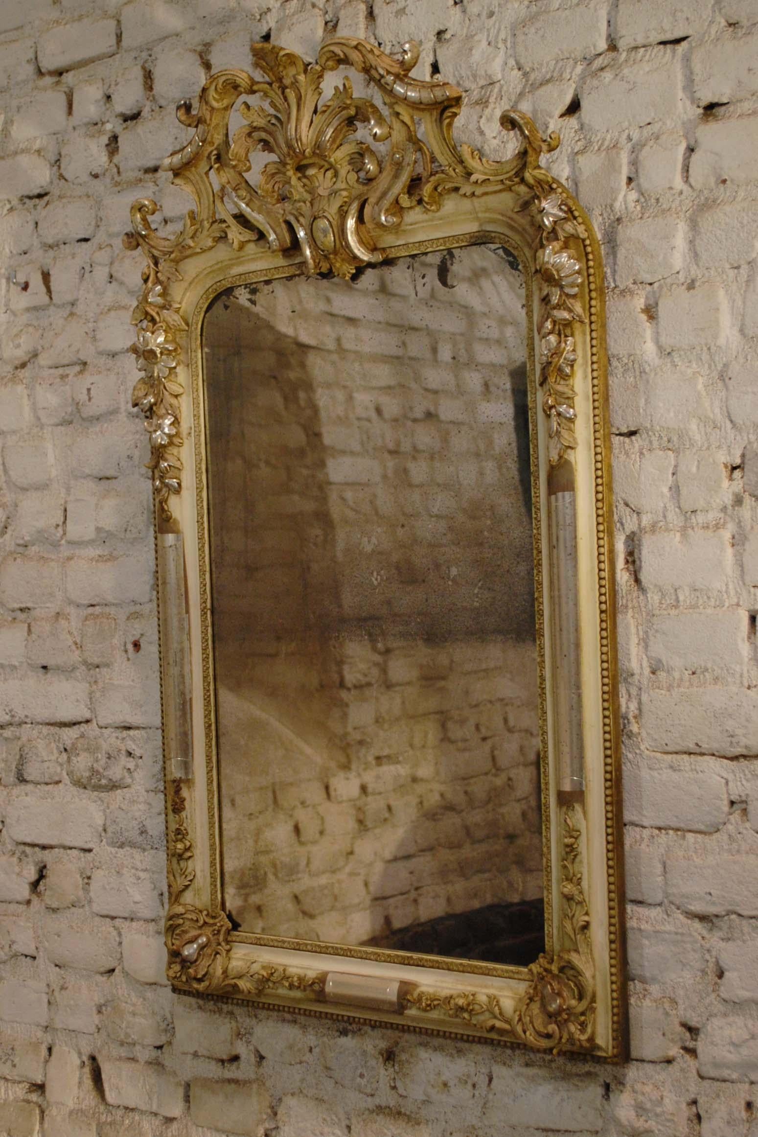 This beautiful mirror is partially silver leaf gilded and shows the bone color of the gesso.
The frame has the upper rounded corners typical for the Louis Philippe style. It features rich ornaments around the frame, such as bay leaf, scallops,