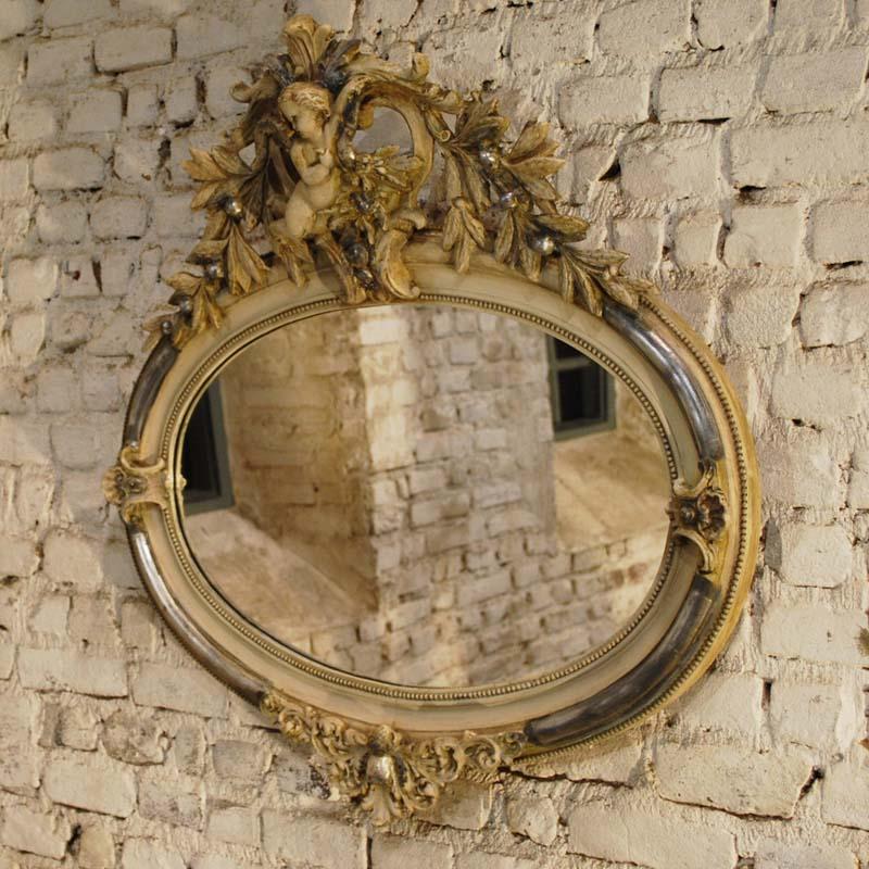 This beautiful oval mirror is partially silver leaf gilt. It has remnants of gold paint and it shows the bone color of the gesso. 
It features rich ornaments around the frame, such as bay leaf, scallops, and a big baby angel or putti. The frame has