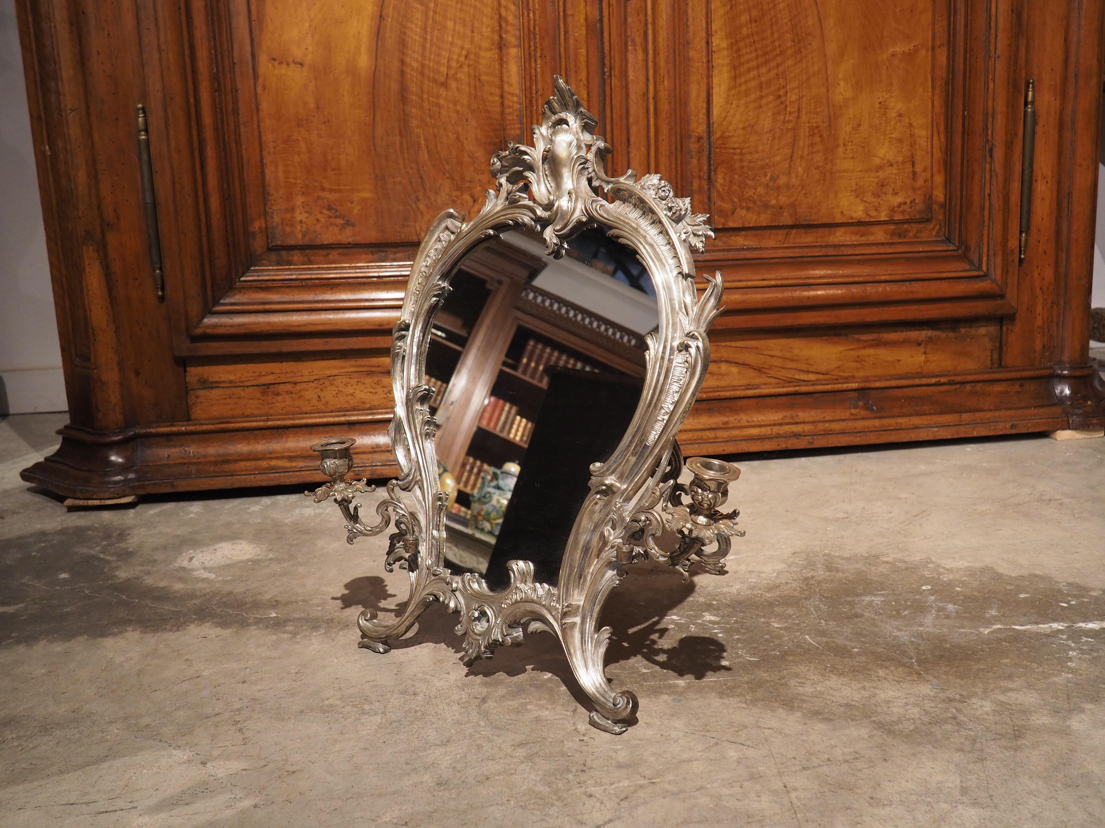 A unique production from the Napoleon III period, this silvered bronze table mirror features two adjustable foliate candle holders that slot into gudgeons affixed near the base of a Rococo style frame. Comprised of pierced and curled leaves,