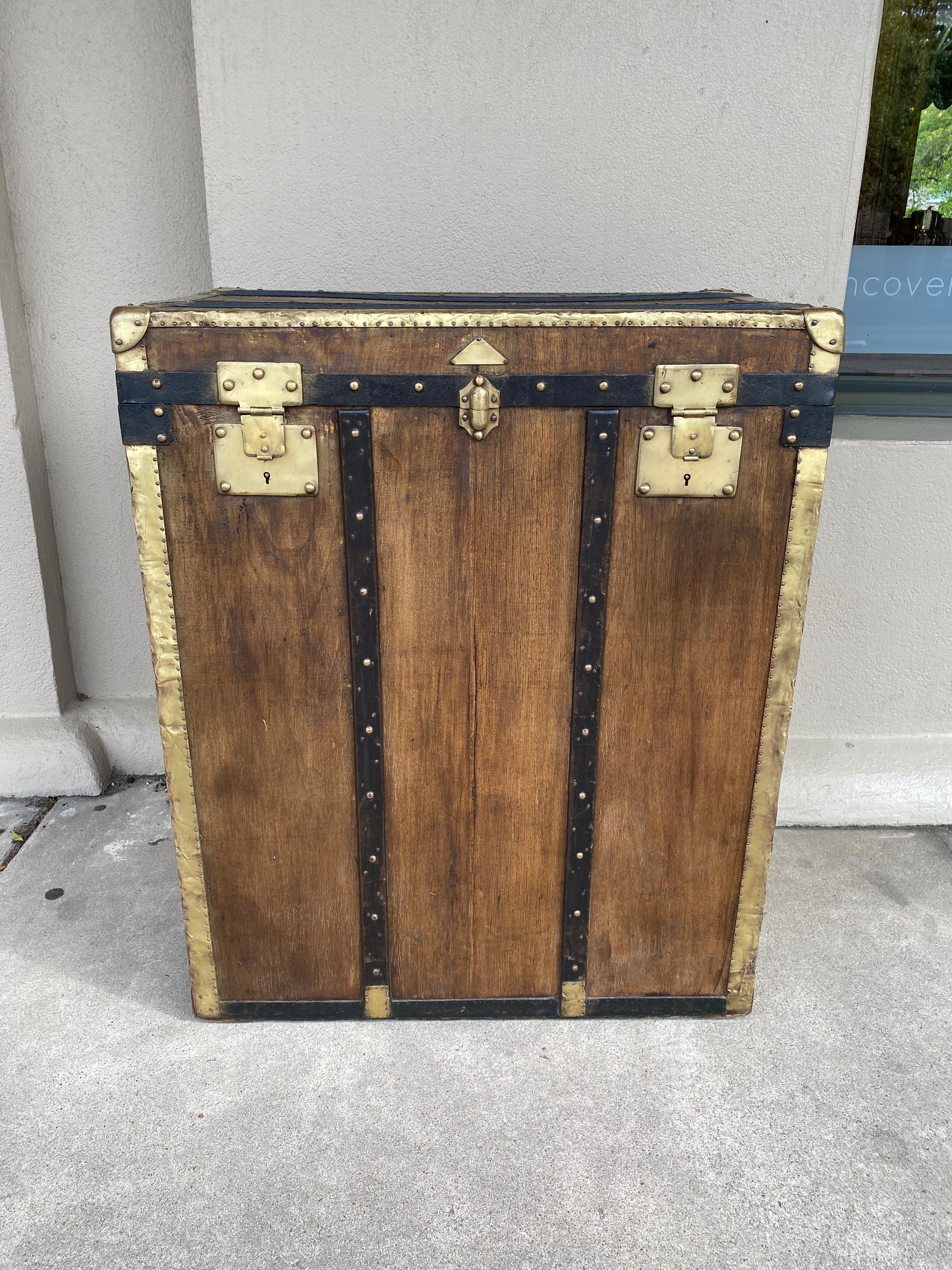 This antique French steamer trunk features a wood body wrapped in canvas and leather with brass hardware and leather handles. The side of this trunk has an old label that reads 
