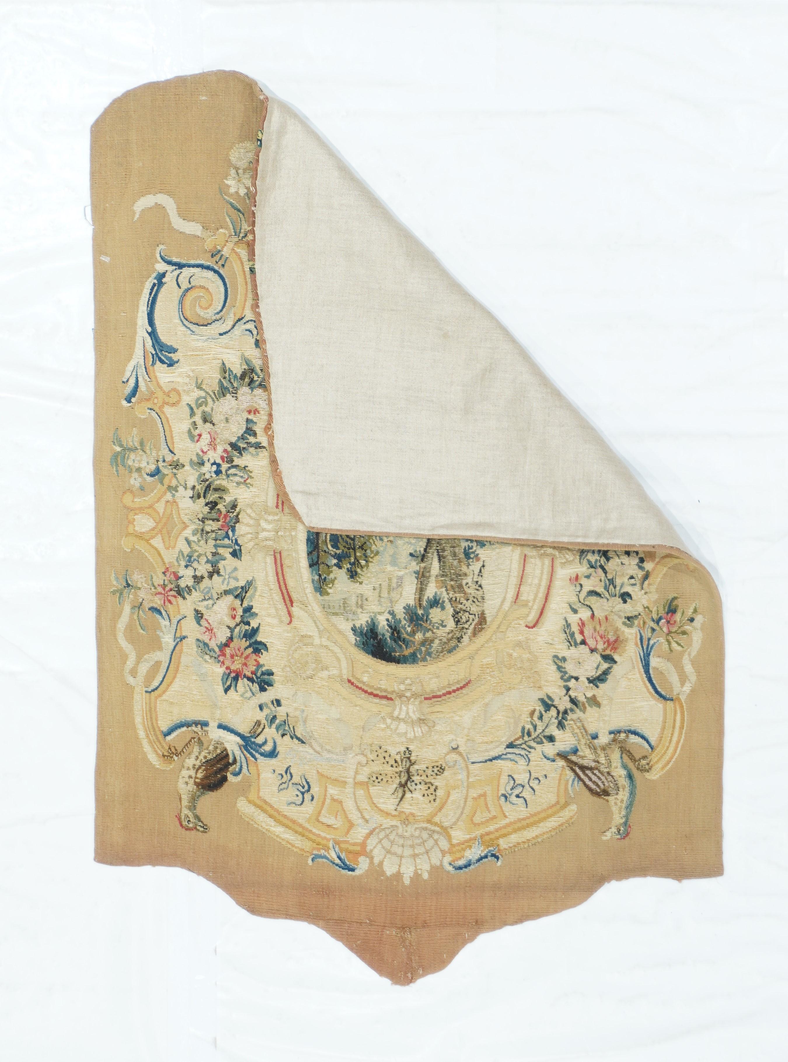 Antique French Tapestry 2' x 3'. The shaped beige field displays an acanthus volute cartouche enclosing an oval floral wreath and an inner rustic image of three tussling dogs beneath a tall, leafy tree A hill is topped at the upper right by a