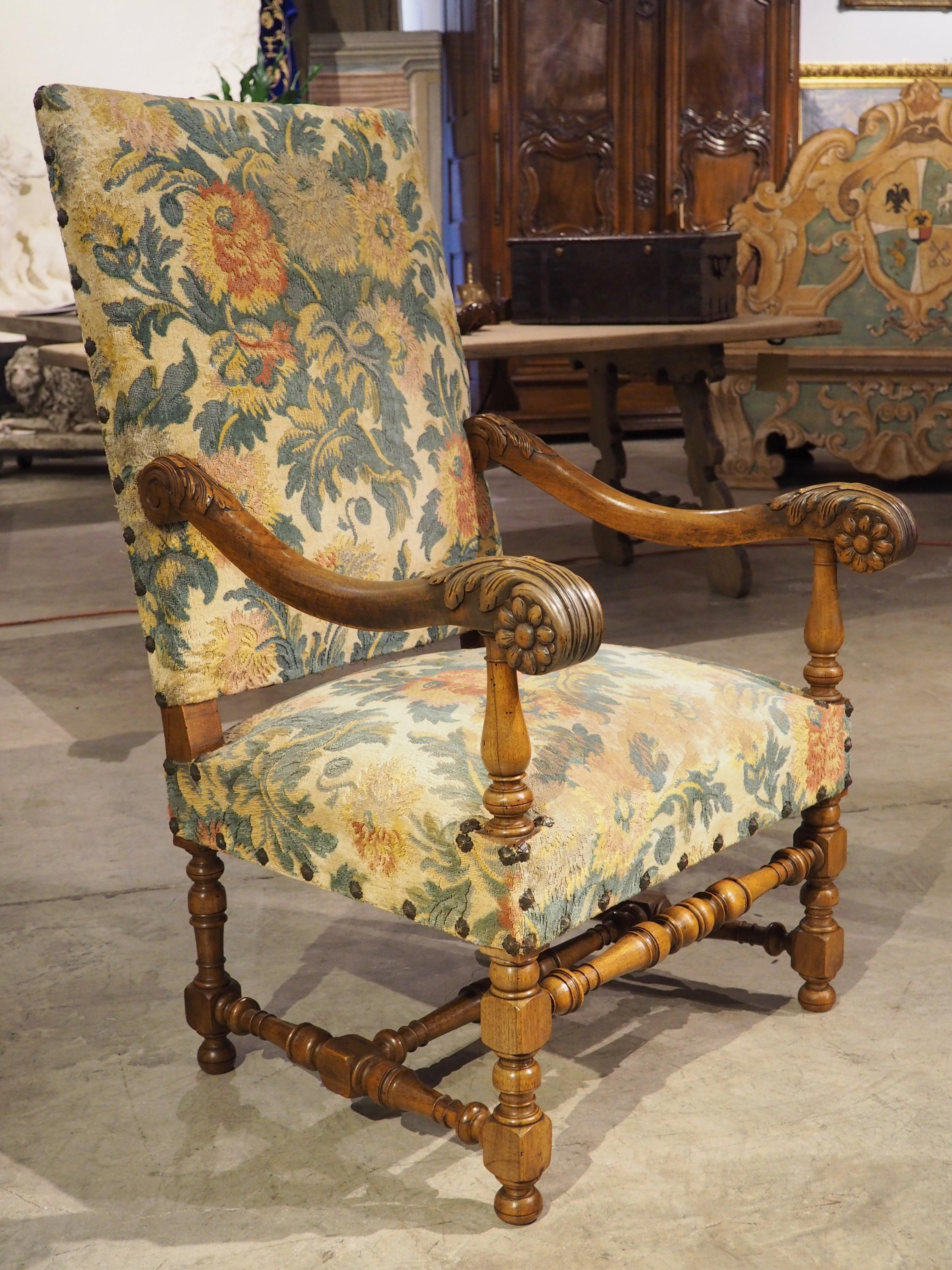 This French walnut fauteuil was hand-carved in France in the 1800’s in the style of Louis XIV. The seat and back have been upholstered in a velvet with green, yellow, and red floral print over a gold background. It is affixed by brass rosettes along