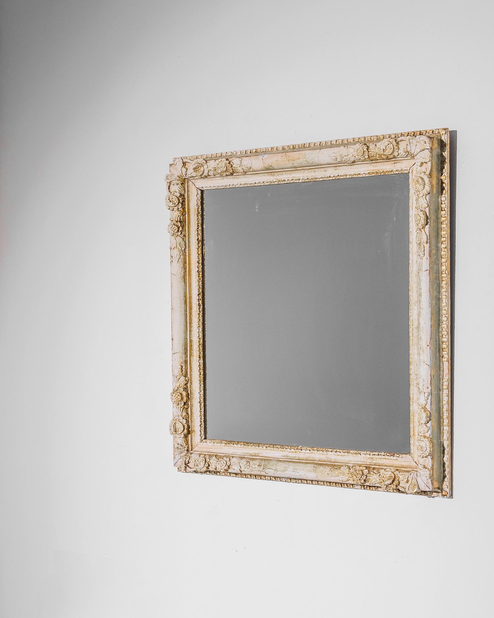 A wooden mirror produced in France circa 1880. This gilt, antique mirror, measuring 47 inches tall, features carved rosette and floral patterns around its deep frame. As a looking glass framed in glittering gold and white, it is meant to be gazed