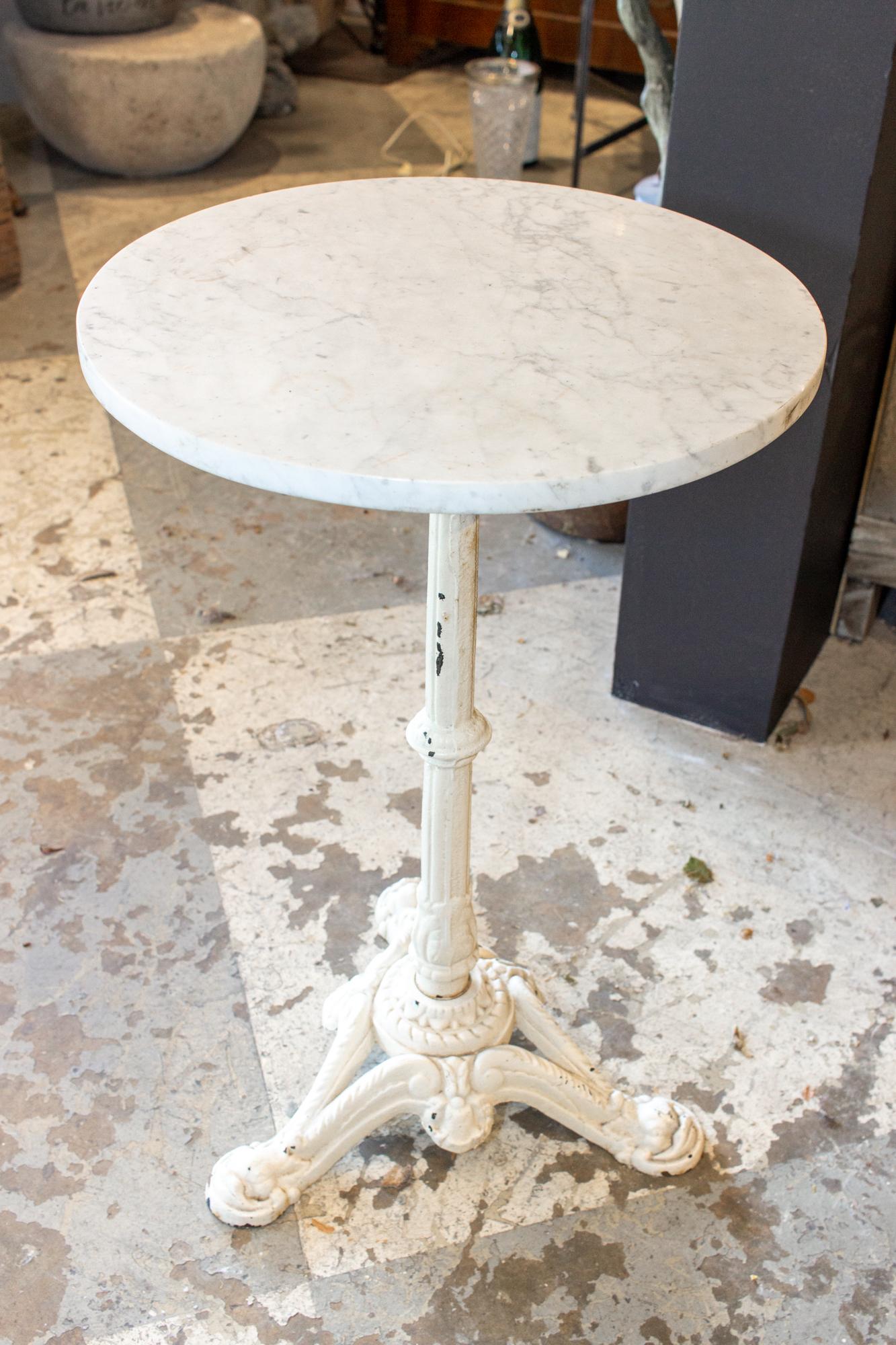 This petite bistro table features a white marble top with grey veining and a beautiful iron base that has been painted white. The top is in good condition with light surface wear consistent with the age and use. The base has some decorative details