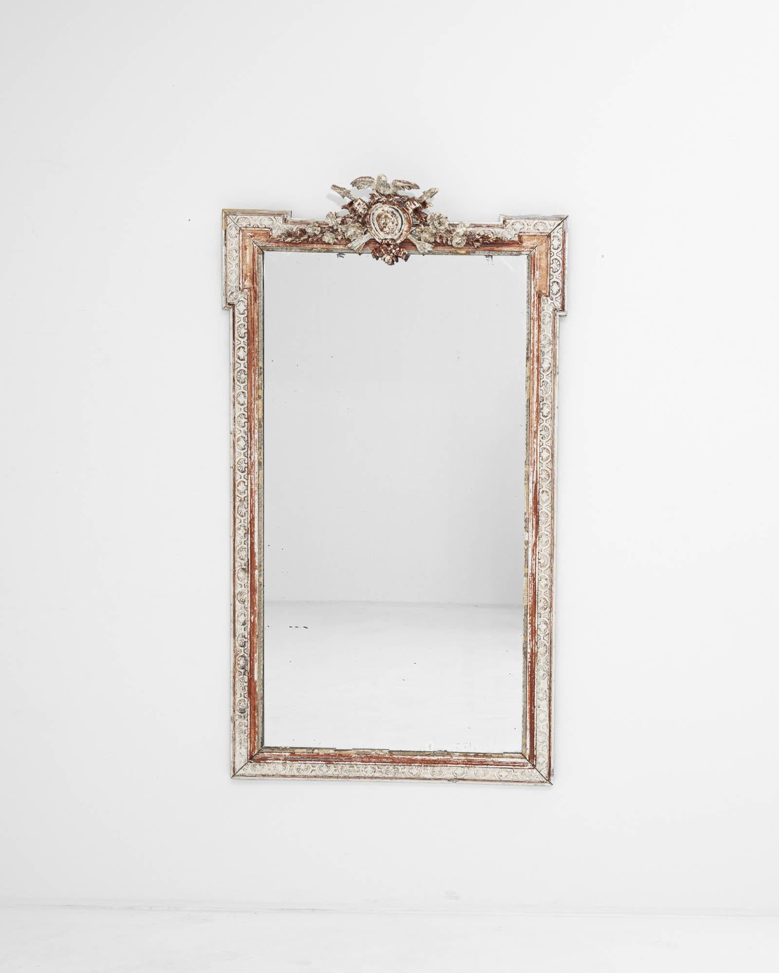 Hand-crafted in the 19th century, this exquisite looking glass flaunts a stunning frame with a distressed white patina that gently accentuates the beautiful carved details, causing the rose hue of the wood to glimmer like gold. The sophisticated