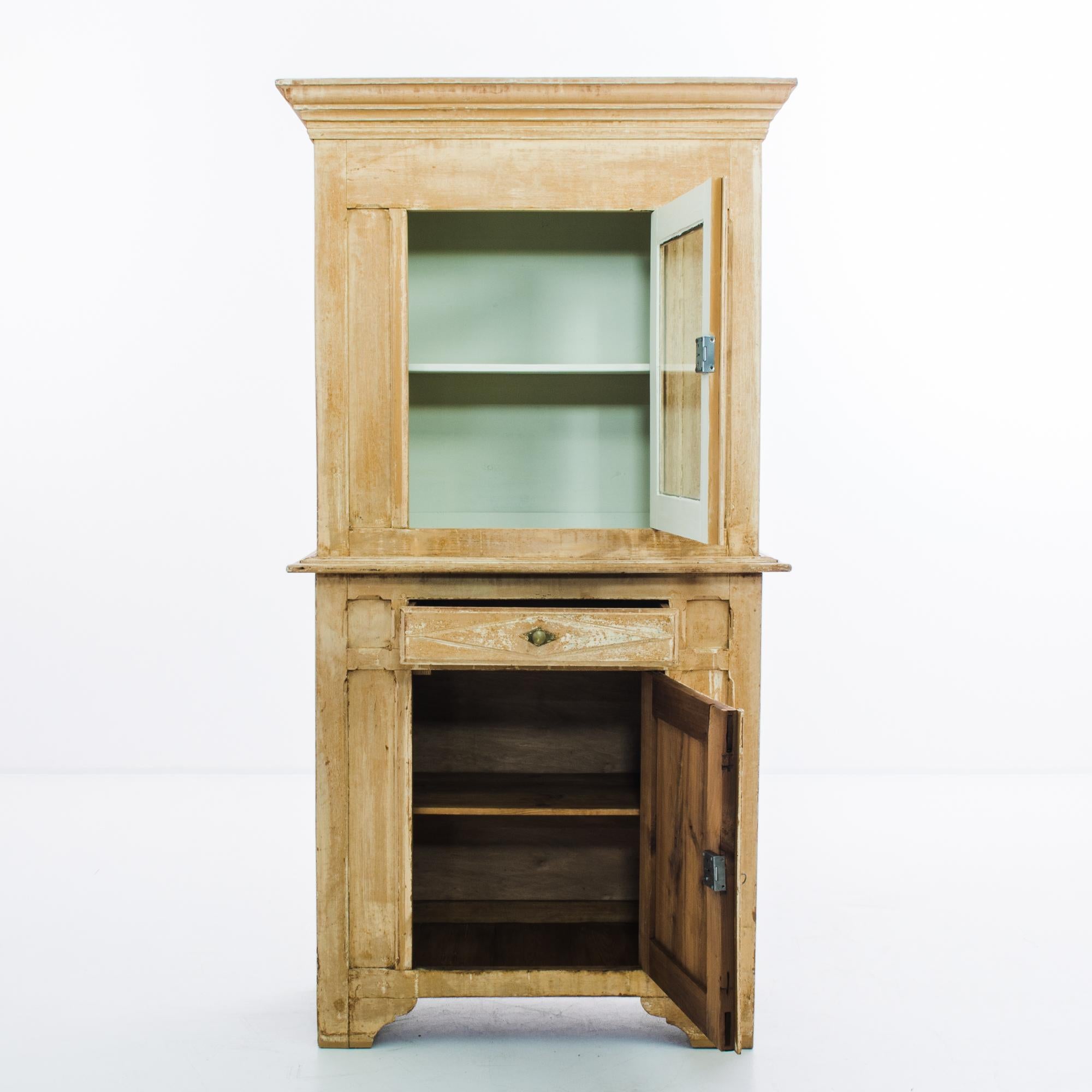 This antique wooden cabinet with a beautifully weathered patina was made in France. It flaunts a rectilinear silhouette, accented by crown molding and bracket feet. The upper vitrine and a lower storage compartment are each fitted with two shelves.