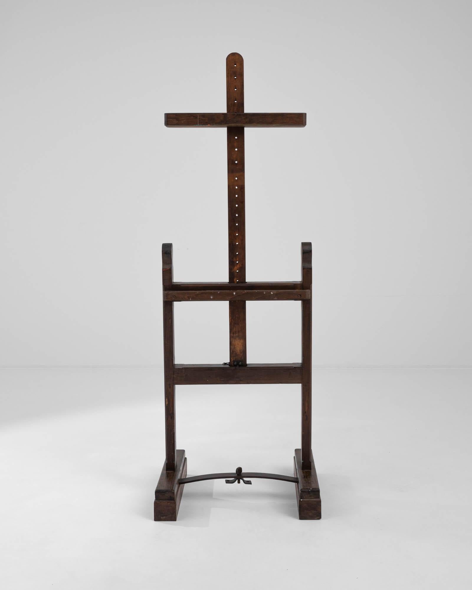 This distinctive cross-shaped easel was crafted from dark, refined wood in France during the 20th century. Featuring a traditional H-frame adorned with carved finials, it stands resolutely on its sturdy rectangular feet, connected by a metal side