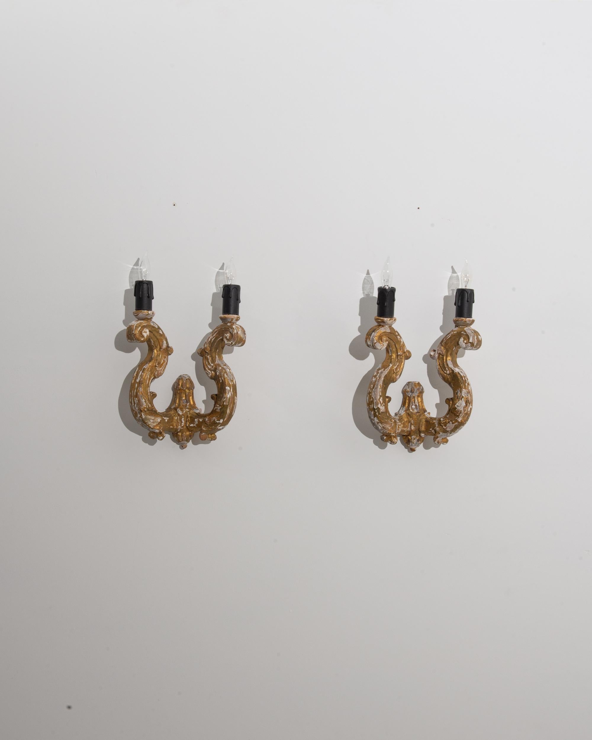 A pair of wooden wall lamps created in 1900s France. These light fixtures are scrupulously carved, forming an enlightened scroll motif that spirals upward to support two symmetrical bulbs. The candelabras of these sconces have been sculpted to seem