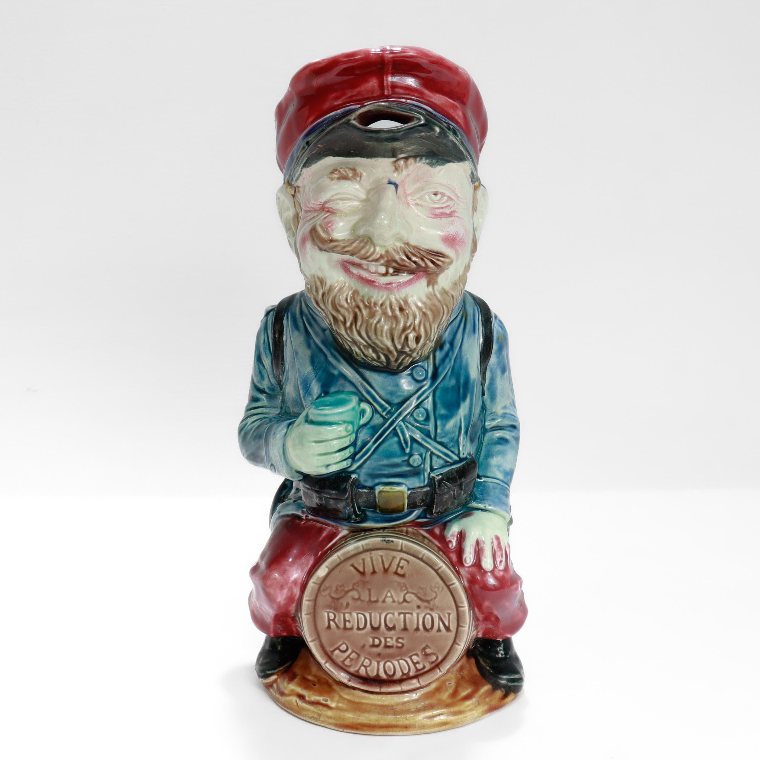 A fine, figural French toby pitcher. 

By Frie Onnaing.

In polychrome decorated majolica.

In the form of a drunken soldier sitting astride a wine barrel inscribed with an anti-establishment slogan “Vive La Reduction des Periodes” for 