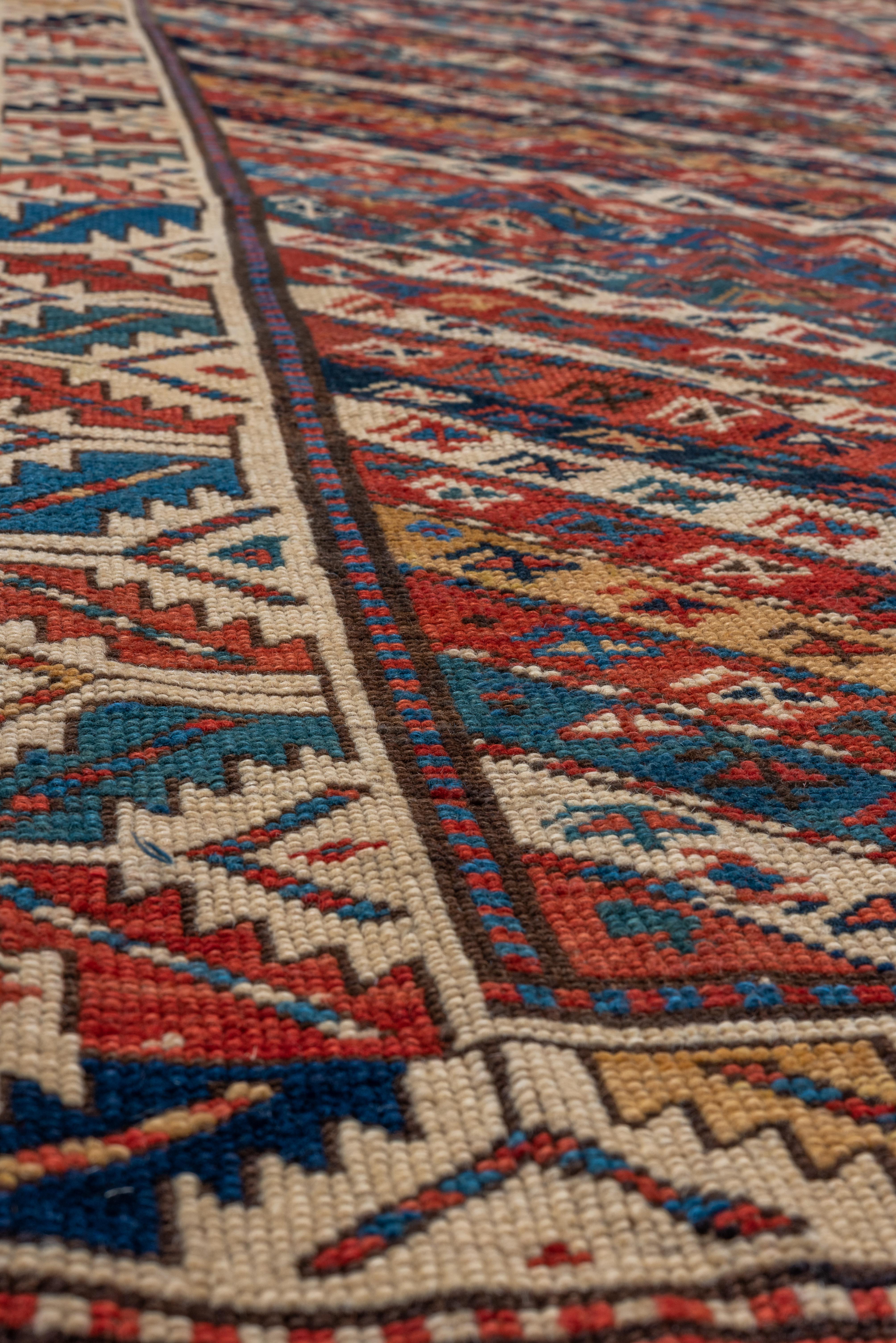 This antique Genje rug showcases a dynamic diagonal stripe field composed of repeating tribal motifs in rich tones of madder red, indigo, ivory, and ochre. Each stripe is filled with geometric icons including stars, hooked diamonds, and S-forms,