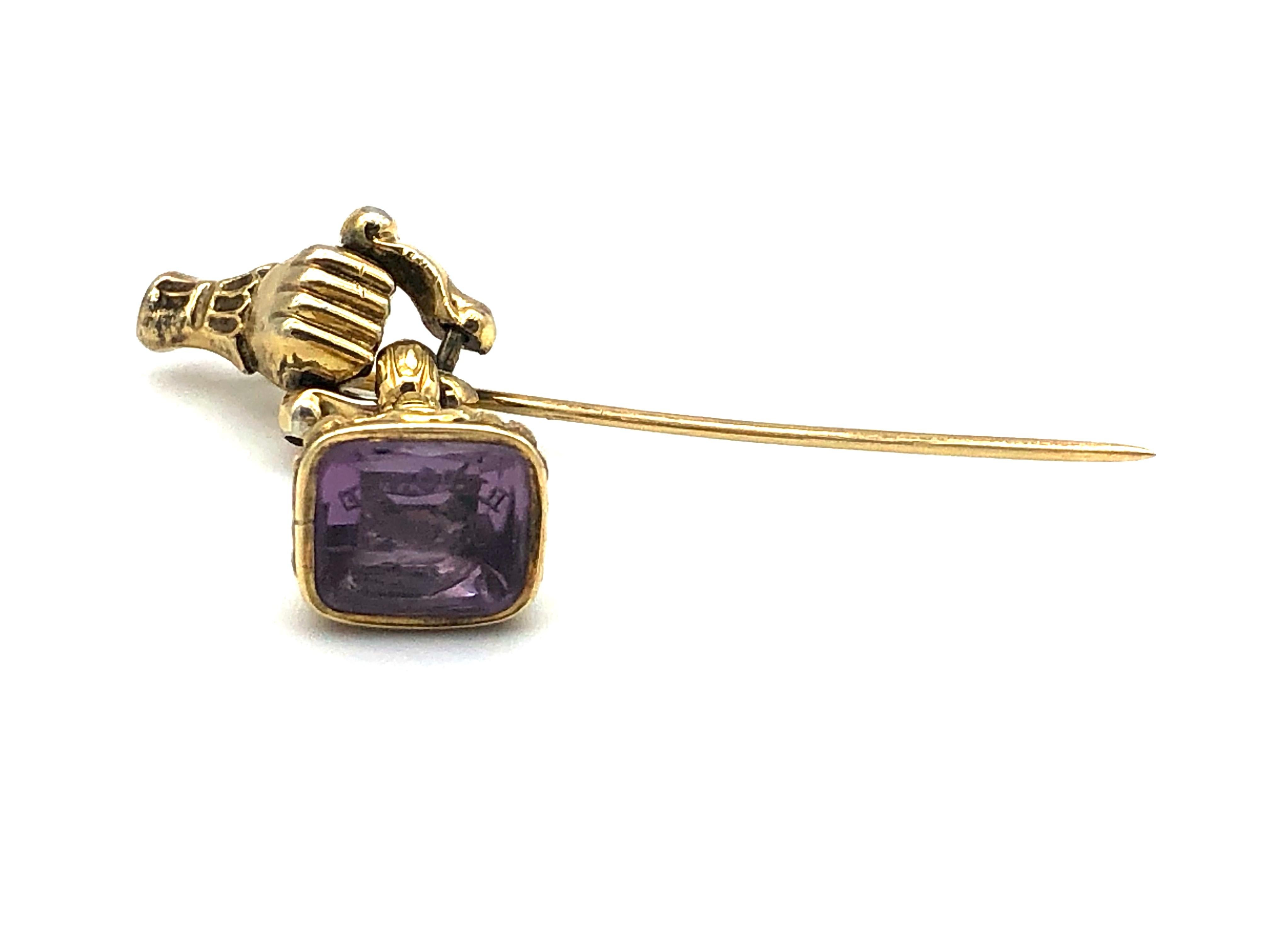 Rare stickpin with a gold hand in a lace cuff. The hand is clenched to a fist and it is holding a hinged element reminiscent of a door knocker in the shape of an ornate stirrup.  A gold seal with an amethyst intaglio is suspended fom this 'stirrup'.
