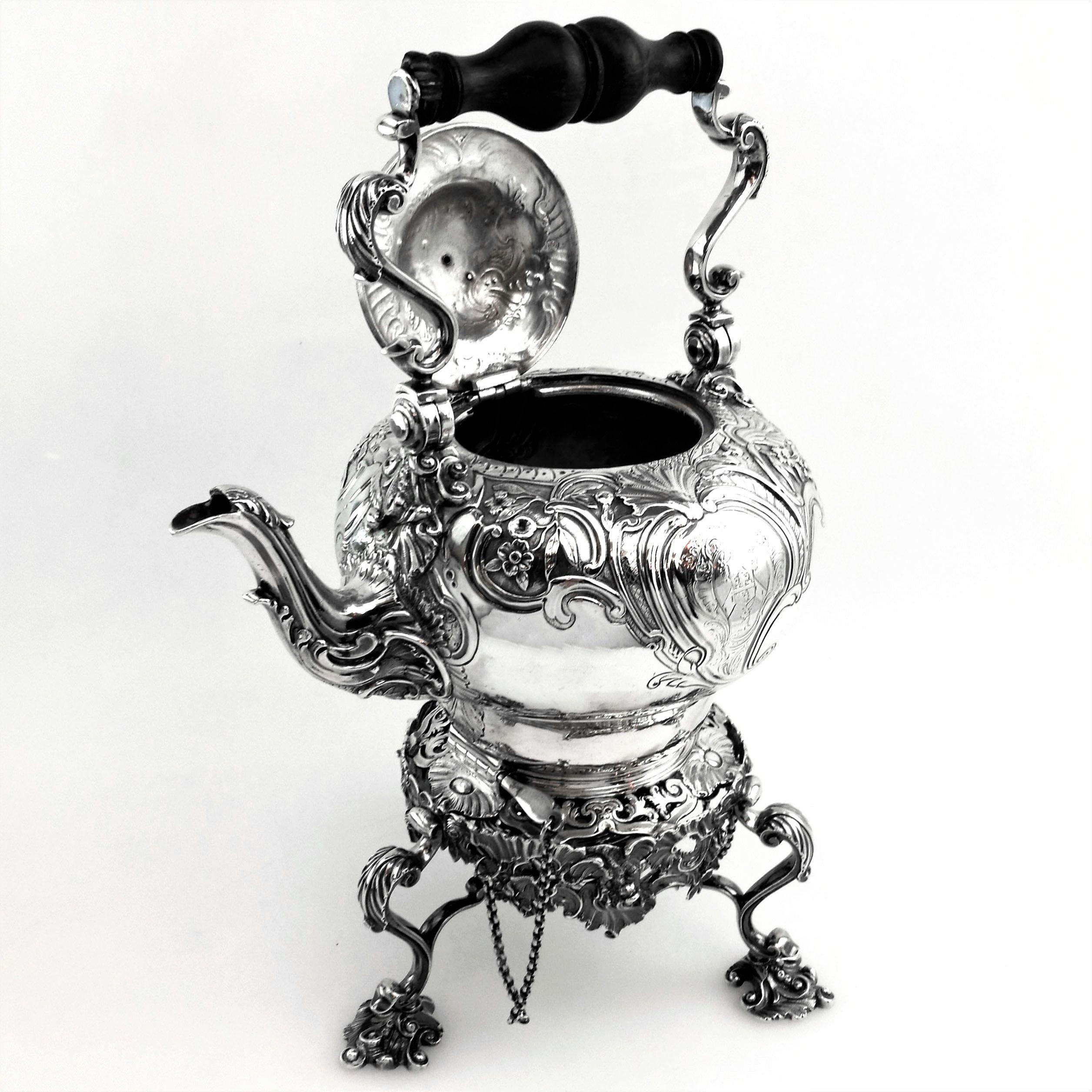 An excellent George II Georgian Solid Silver Kettle on Stand. The Tea Pot has an ornate chased design including floral, foliate and scroll patterning, and each side has a shaped cartouche with an engraved crest in it. The Teapot has a hinged domed