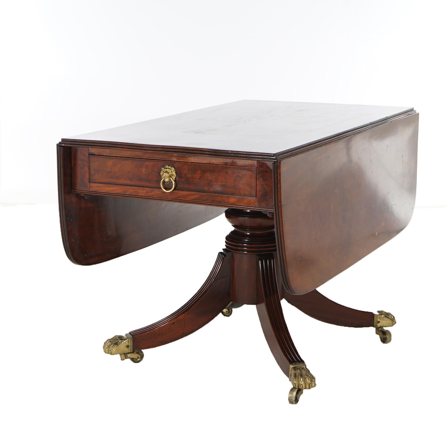An antique English George III style table offers mahogany construction with top having drop leaves over single drawer having flame mahogany facing and cast lion head pull surmounting urn form pedestal base having four Duncan Phyfe reeded legs