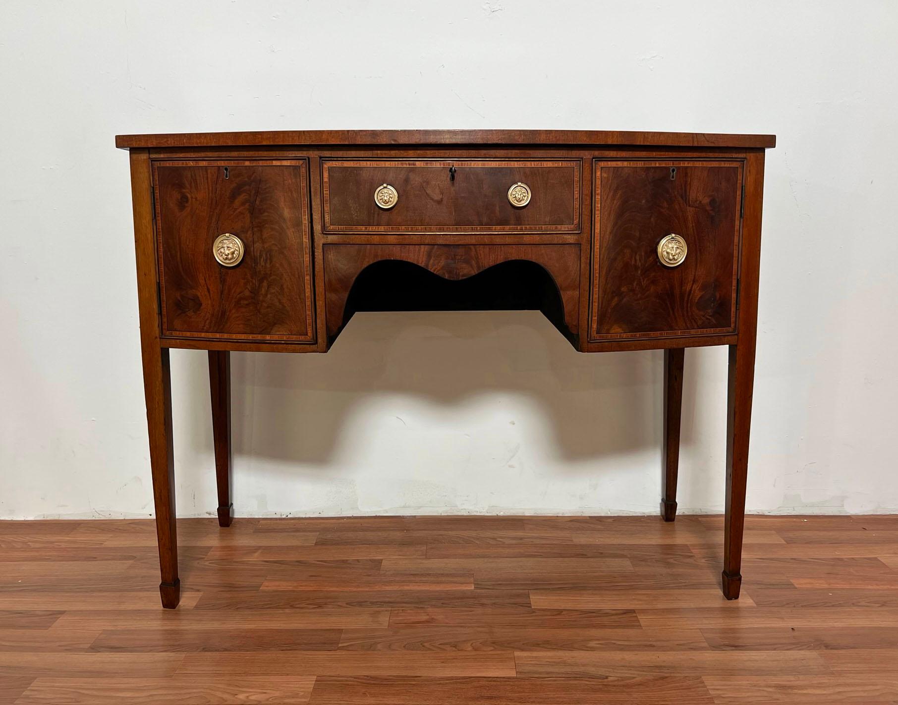 An antique George III era Hepplewhite bow front dressing table, ca. 1810,. Mahogany with contrasting boxwood banding and original rondelle lion head brasses. Also includes original working key.