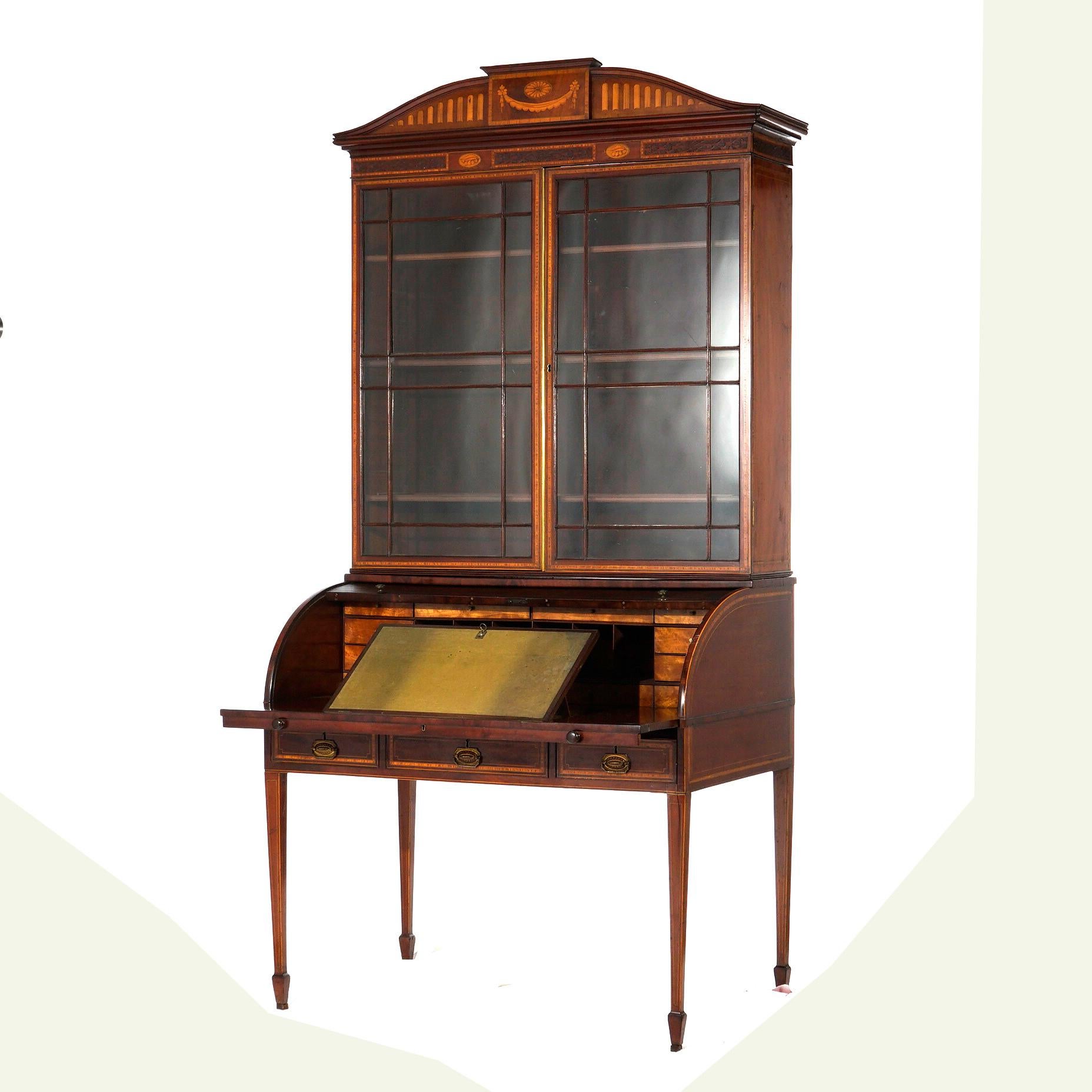 An antique English George III Hepplewhite secretary offers mahogany construction with architectural crest having satinwood inlaid elements over double glass door bookcase surmounting roll top desk with lower drawers, raised on tapered straight legs