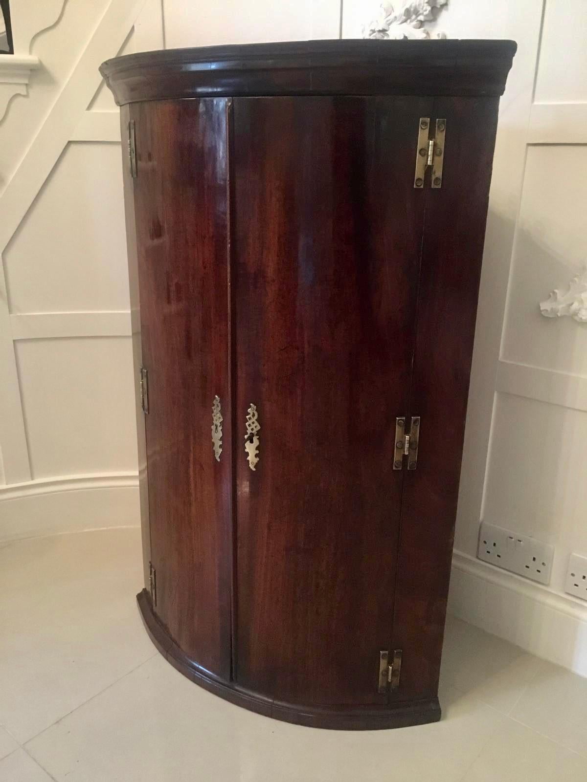 Antique George III mahogany hanging bow front corner cabinet with six original H shaped brass hinges and original brass escutcheon. It boasts a pretty shaped moulded ogee cornice and two beautiful mahogany bow front doors which open to reveal 3