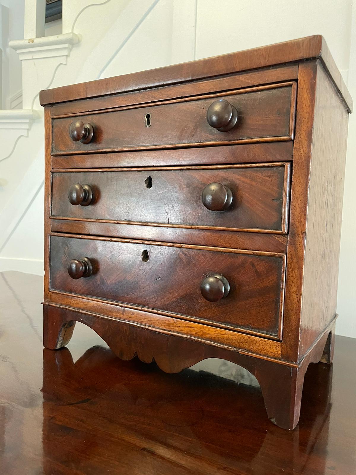 Antique George III quality figured mahogany miniature chest of 3 drawers having a quality crossbanded figured mahogany top above 3 mahogany cockbeeded drawers with original turned knobs standing on splayed bracket feet united by a shaped apron 

A