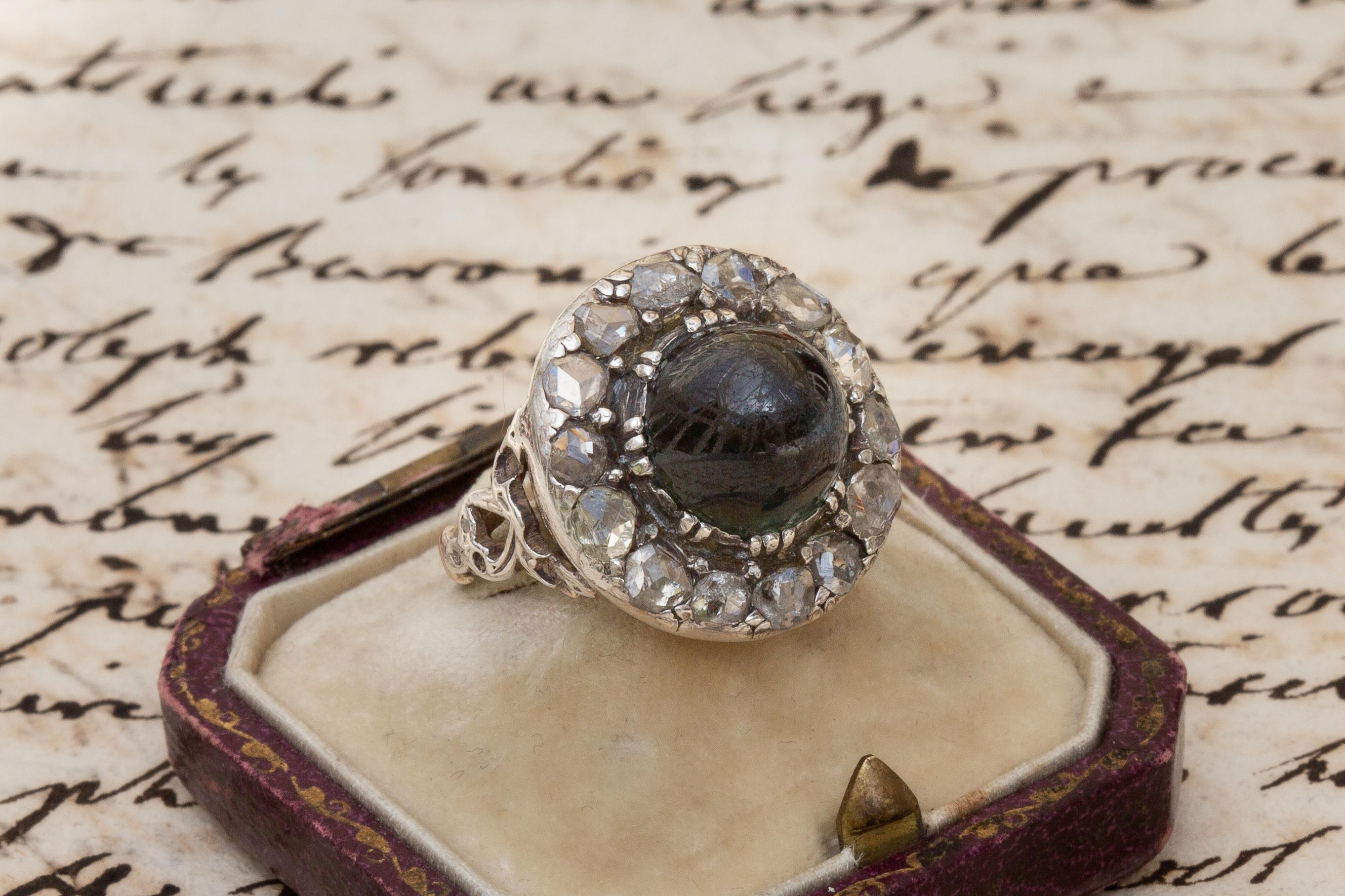 This unusual cluster ring dates to the the early 19th century, circa 1810. A rare and usual example, this Georgian era cluster is set with a high-domed bi-colour sapphire which displays a predominant deep chromium green colour with areas of teal