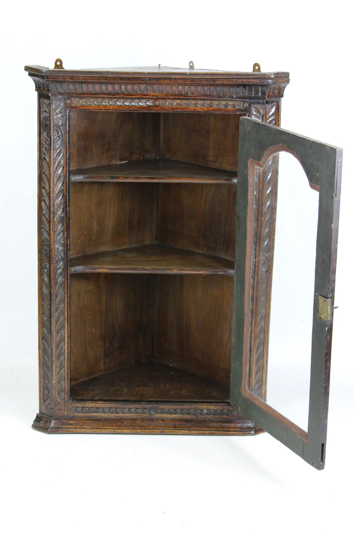 An attractive small antique Georgian hanging corner cupboard in extensively carved oak with a single glazed panel door. It has a working lock and key (replacement) and 2 shelves to the interior. It has a fancy brass escutcheon and brass H-hinges to