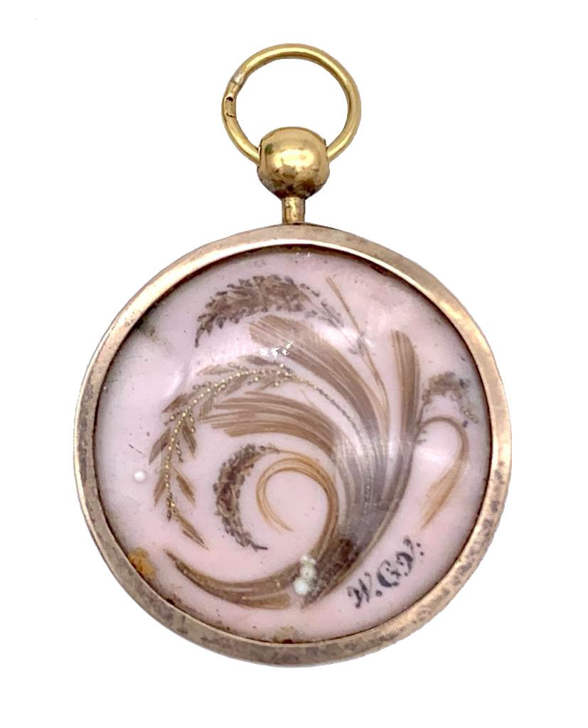 This delicate mourning pendant features beautiful work on both sides. Round porcelain plaques are painted and decorated with hair. One side shows a large urn carrying the initials JMS on it's base in front of a weeping willow. The lid of the urn is