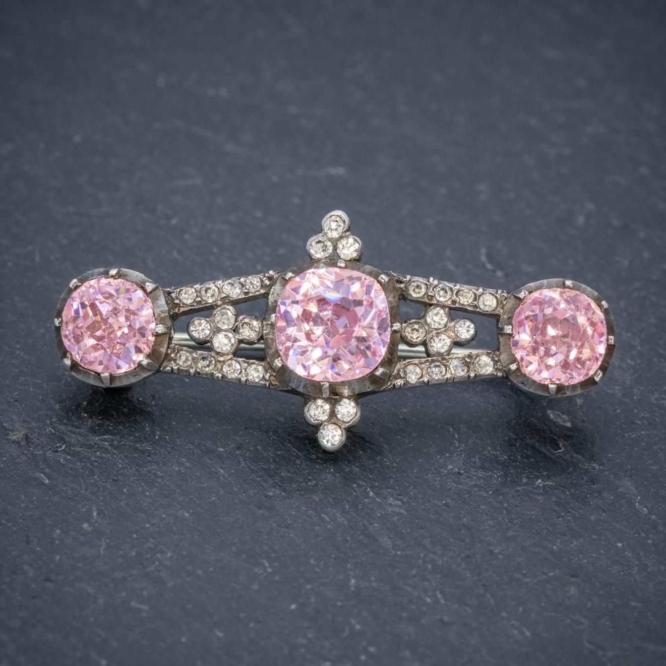 A lovely antique Georgian bar brooch crowned with a trilogy of shimmering pink Paste Stones the largest of which is 2ct in the centre flanked by smaller 1.20ct stones. 

The gallery is modelled in Silver and dotted with smaller white Pastes which