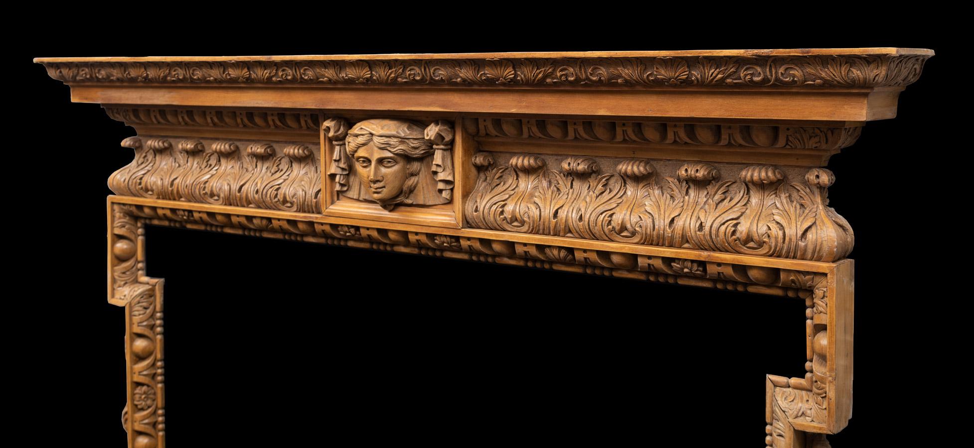 A tall and robustly carved George II style pine chimneypiece in the manner of William Kent. The acanthus decorated frieze with female mask at centre is under an egg and dart cornice shelf. The side scrolled consoles carved with acanthus, acorns and