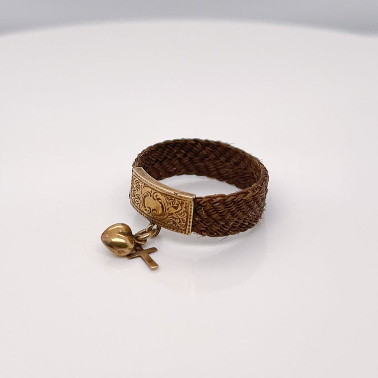 A fine antique Victorian or Georgian mourning ring.

Made primarily of woven hair with an engraved gold band and two diminutive charms.

The band is engraved with floral motifs and holds two small charms - one in the shape of a cross, the other a