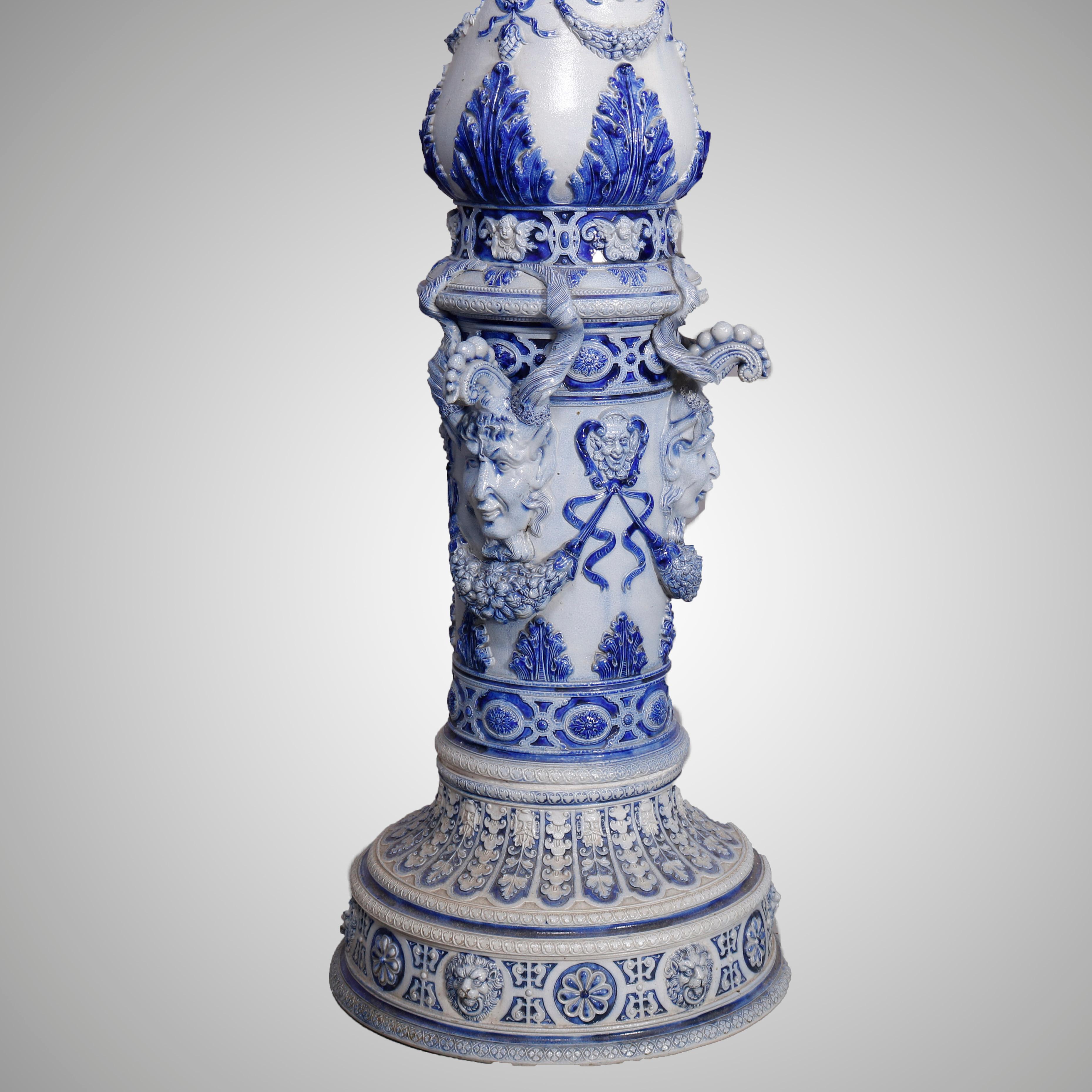 An antique German stoneware jardinière and pedestal offer blue and white stoneware construction with urn form planter offers high relief neoclassical scenes including cherubs, female masks and foliate elements flanked by figural handles and raised