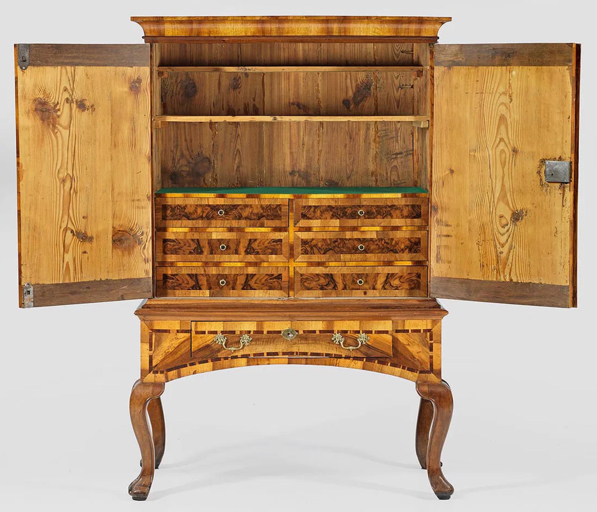 Antico mobile tedesco su cavalletto in impiallacciatura di noce con intarsi in legno di plumbeo. Struttura dritta a un cassetto su gambe fortemente arcuate. Sopra, un piano a due ante con cornice scanalata e modanata. All'interno, scaffali e