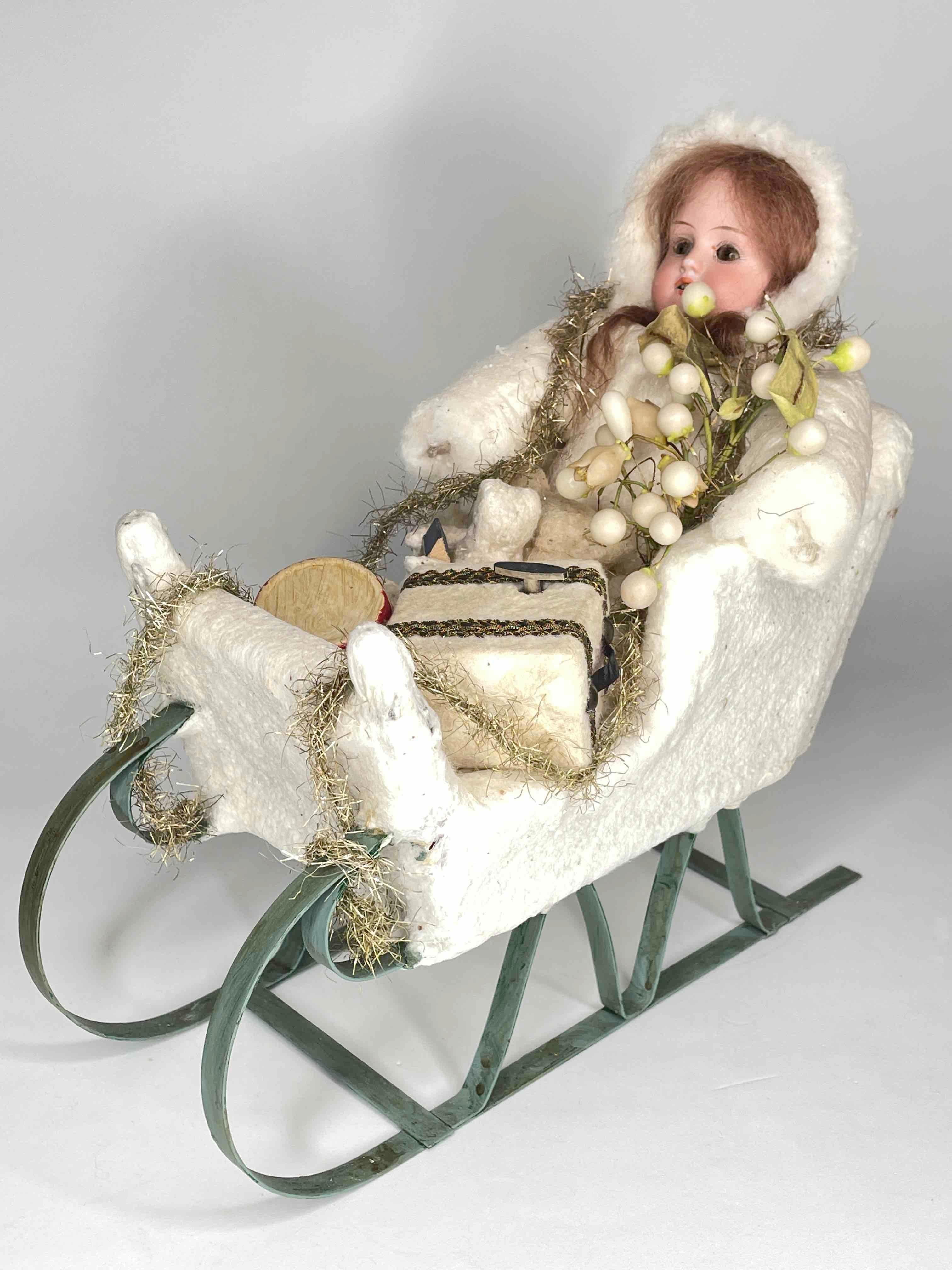 Cette magnifique poupée en biscuit, accompagnée d'un bébé plus petit, est assise dans un traîneau en métal. La poupée a des yeux de verre bruns, des cheveux et des dents blonds. Le manteau est en coton pressé. Le traîneau était bordé de coton pressé