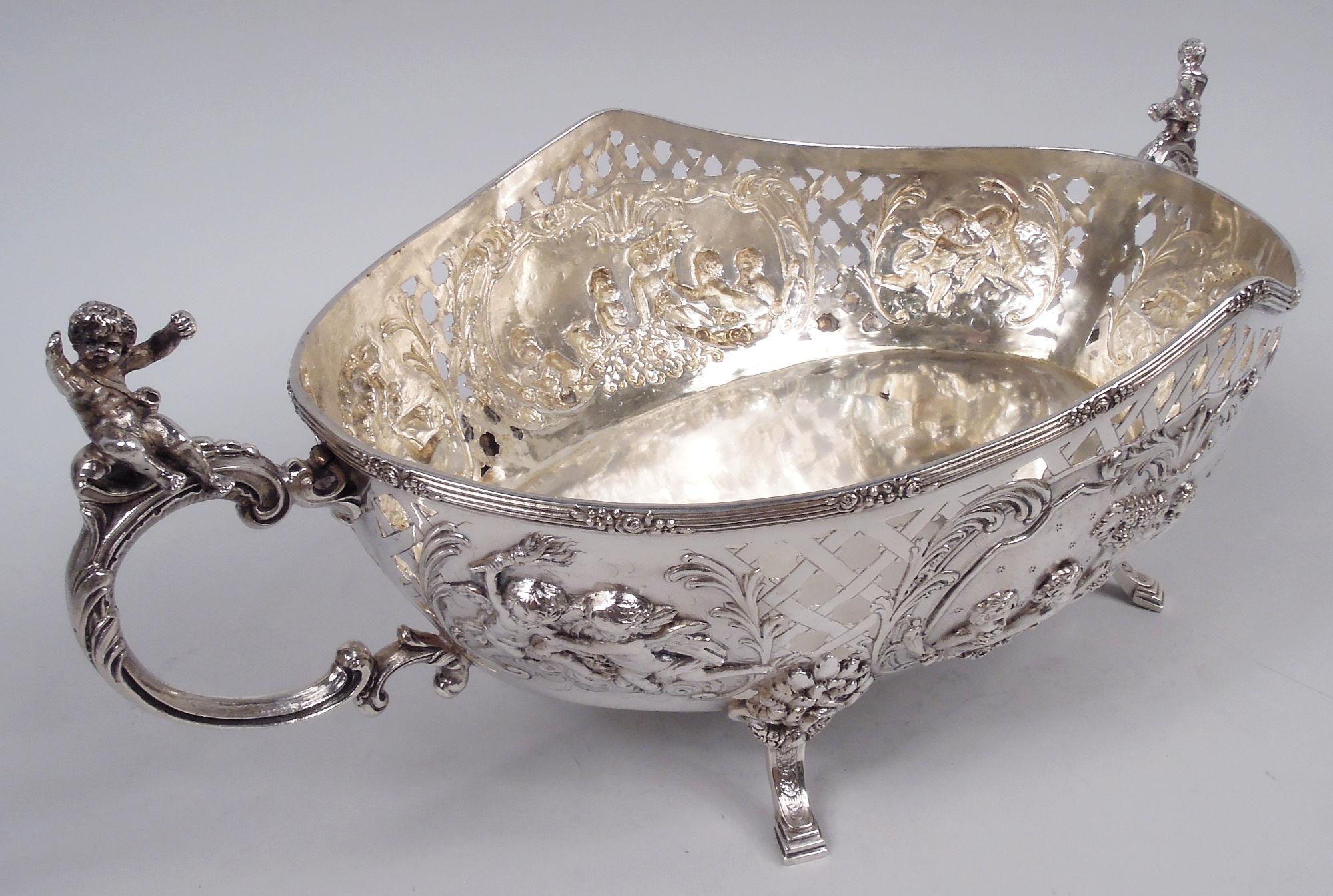 German Classical 800 silver centerpiece, ca 1910. Solid oval well and curved sides with reeded rim, open basketweave, and chased and engraved ornament: Leafing scroll frames inhabited by cherubs feasting on grapes in company of a goat as well as