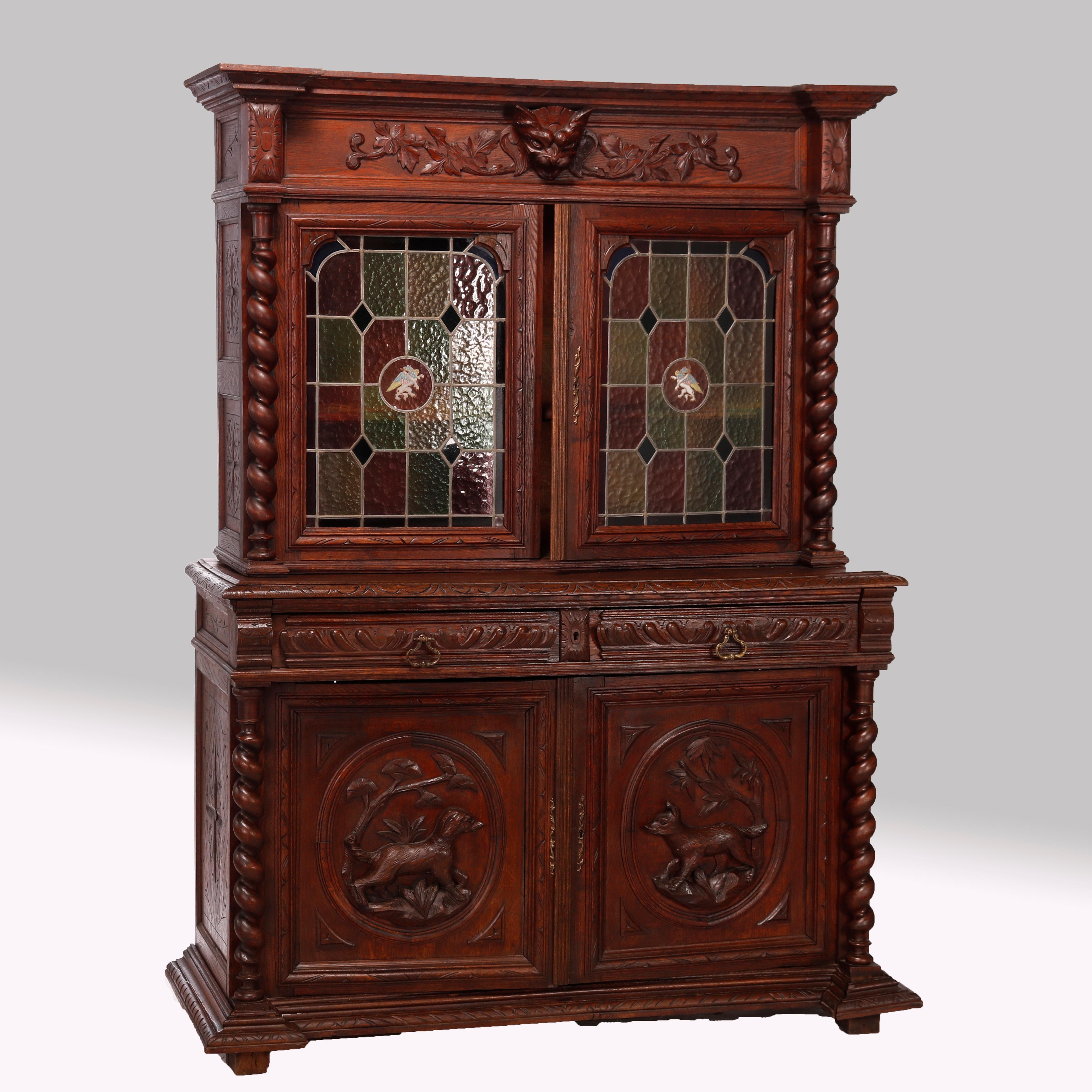 An antique German figural hunt cupboard in the manner of Black Forest, offers oak construction with upper having carved figural mask with flanking foliate elements over double leaded glass door cabinet with central enameled phoenix, flanking rope