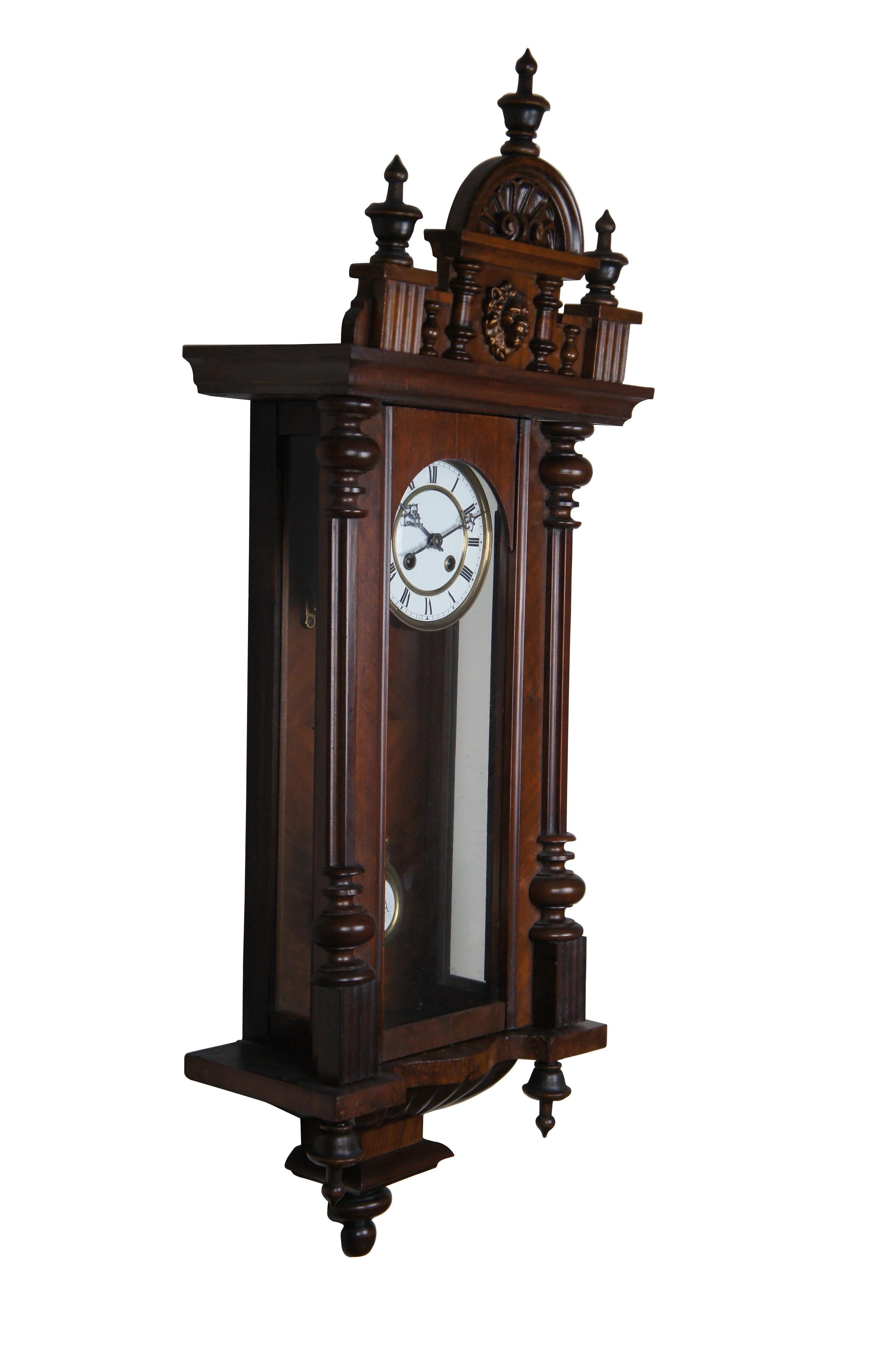 1920s German Mauthe Wall Clock. Features a tall case made from walnut with turned columns, ornate finials and a lion head at the center. The clock face is designed with Roman Numerals and ornate hands. The pendulum is long with at Return and Advance
