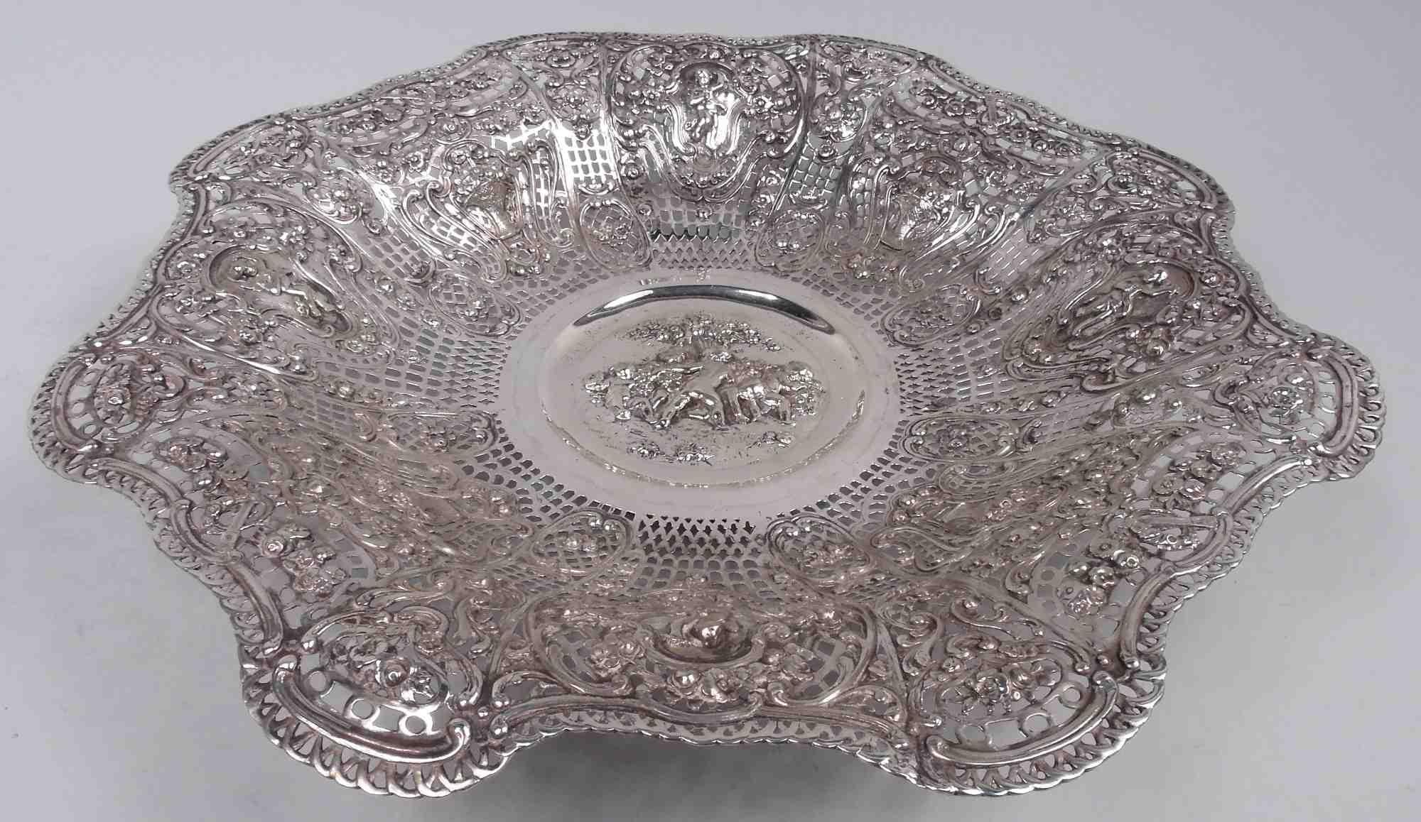 German Rococo 830 silver bowl, ca 1900. Round well with rosebud-gathering cherubs in relief. Sides tapering with scrolled and wavy rim. Heraldic shields with more cherubs and flower baskets as well as flower-strewn scrolled frames on pierced ground.