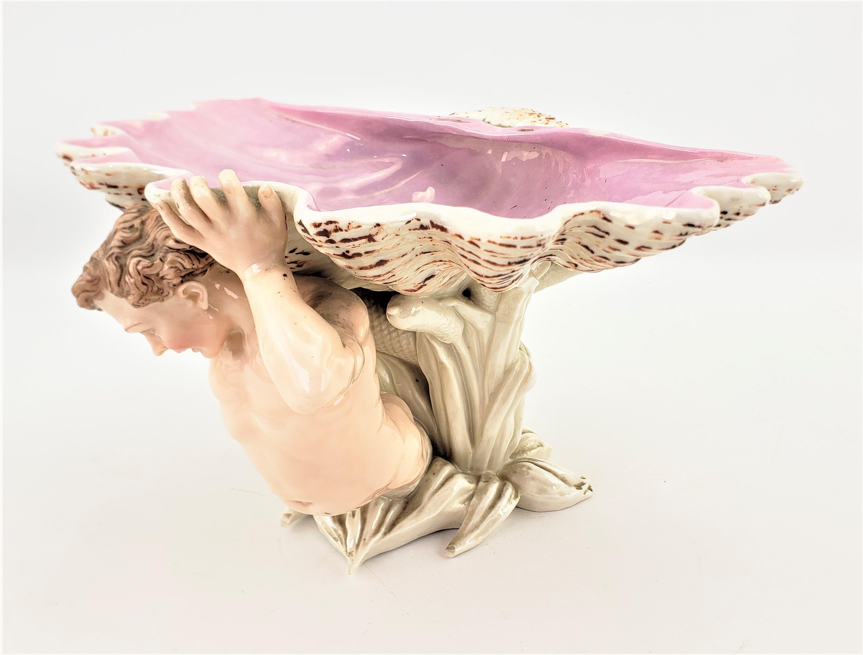 This antique hard paste porcelain bowl or centerpiece is unsigned, but presumed to have been made in German in approximately 1880 in a Neoclassical Revival style. The bowl features a young male mermaid styled figure holding a large shell with a