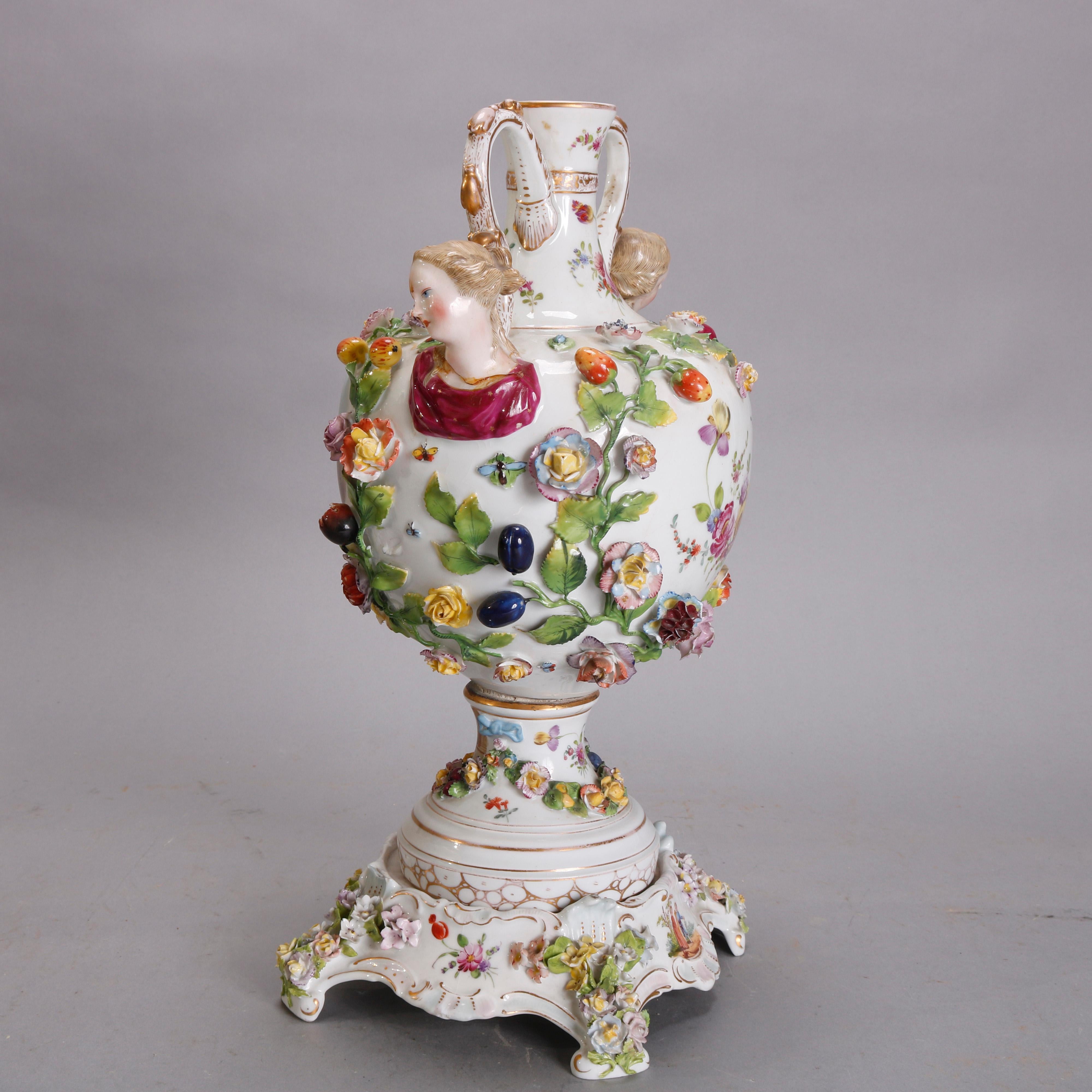 An antique German Meissen urn offers hand painted porcelain construction with central courting scene reserve, double figural bust handles, applied flowers, and gilt highlights throughout, raised on matching decorated porcelain base, blue maker mark