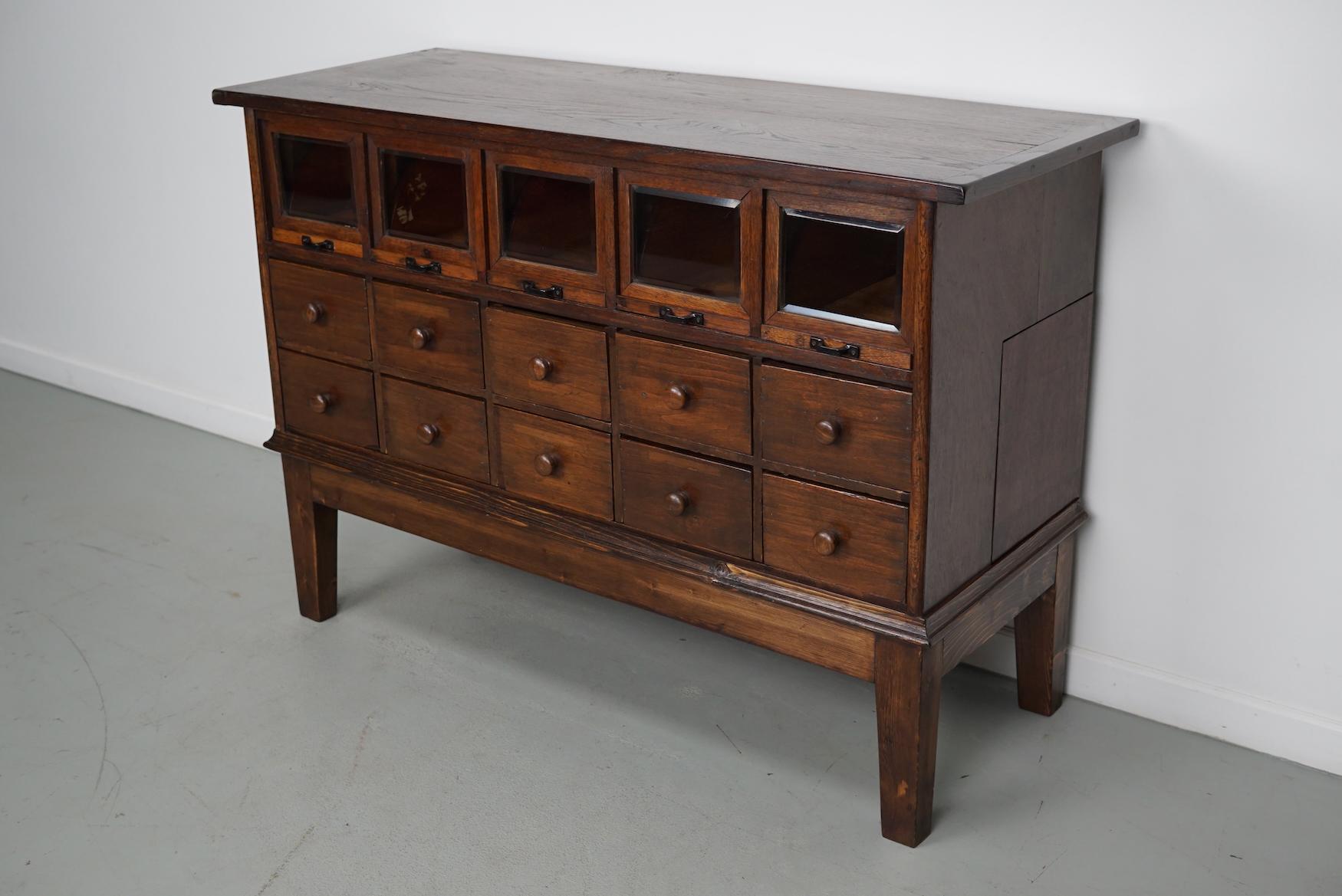This very unique haberdashery cabinet was produced during the 1920/30s in Germany. It features 10 drawers and 5 with cut glass fronts / folding doors and black metal handles and one hidden large drawer in the side of the cabinet. It was originally