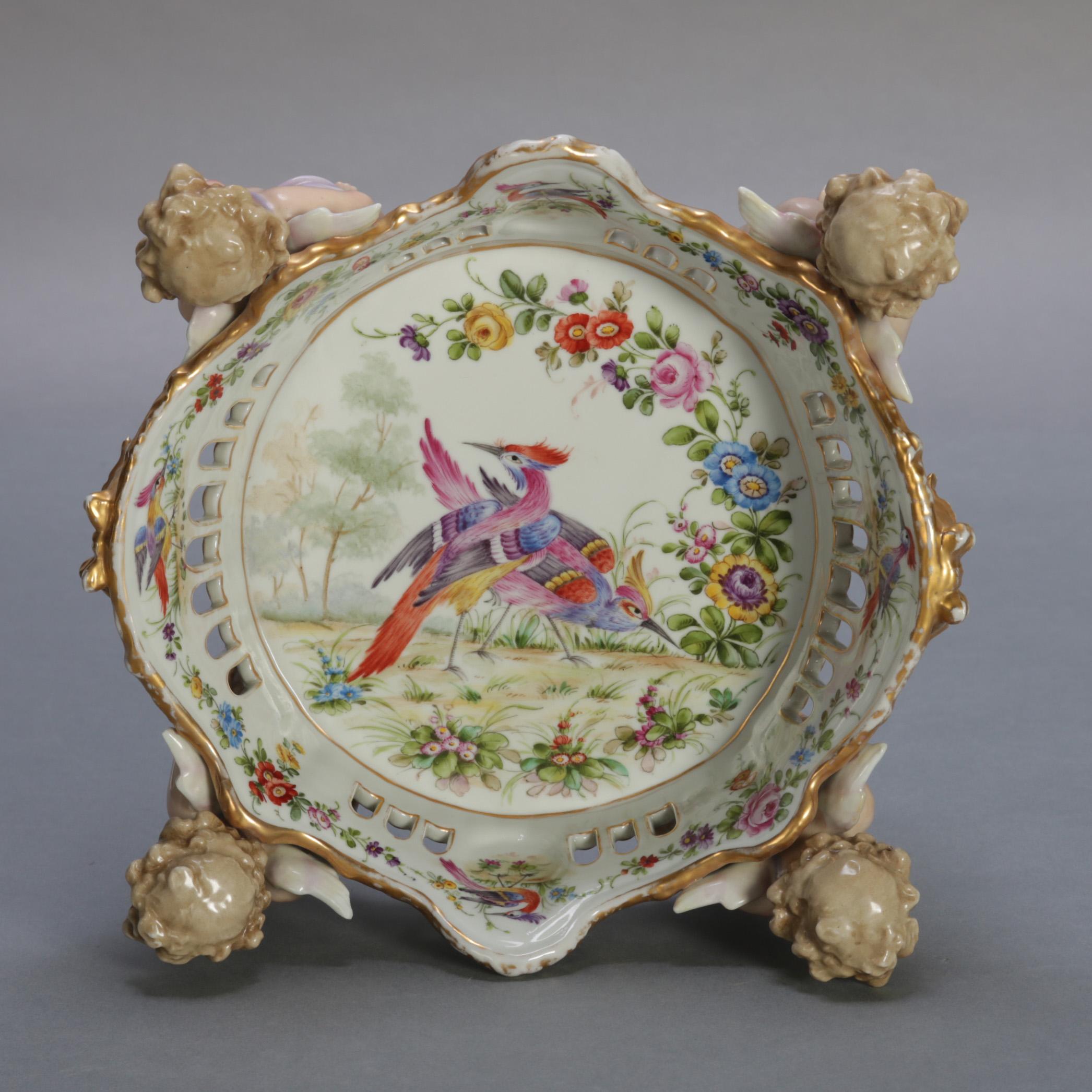 An antique German figural porcelain candy bowl by C.G. Schierholtz & Sohn features four winged cherubi supporting hand painted centre pierced crown form bowl having interior garden scene with exotic birds and all-over floral and gilt decoration,