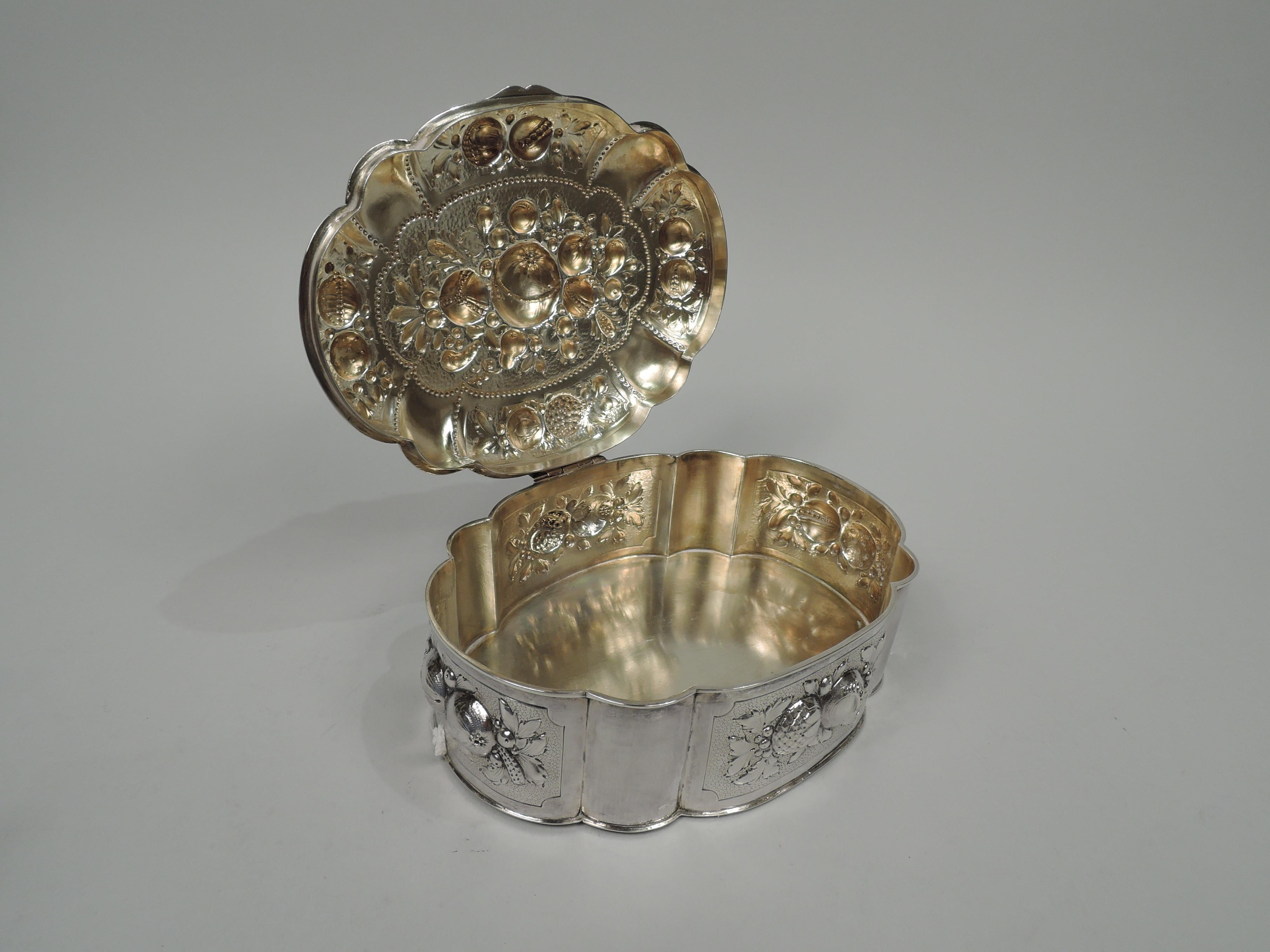 German 800 silver box, ca 1910. Oval and lobed. Cover hinged and raised with scroll tab. Sides have chased and engraved fruits and foliage on stippled ground in curvilinear frames alternating with plain lobes. Cover top same with beaded frames.