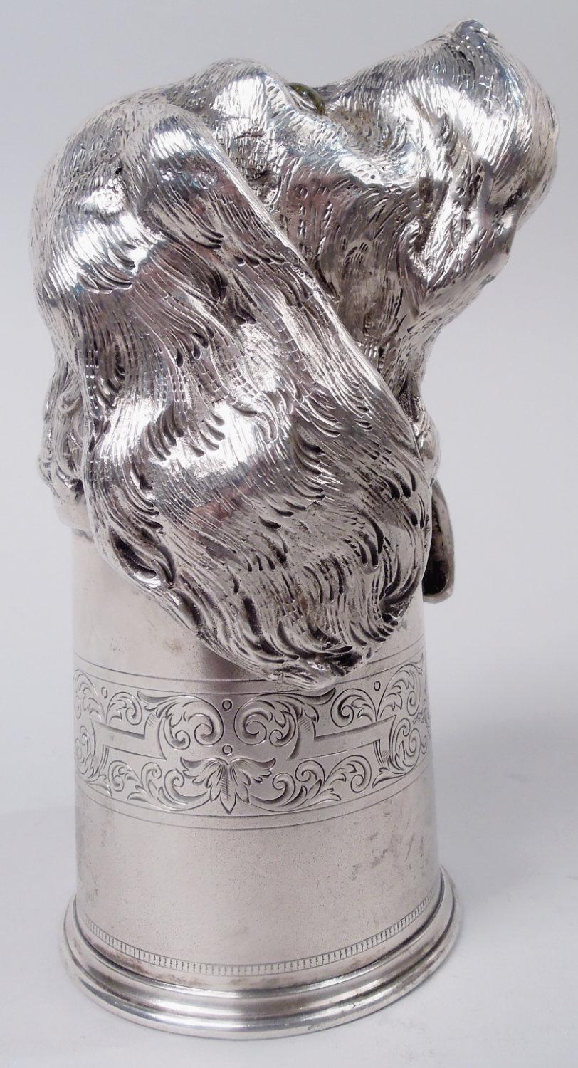 German 800 silver stirrup cup, ca 1930. Cast dog head in form of floppy-eared hound with jowly, upturned snout, and glass yellow eyes with dazed pinpoint pupils. Bowl has straight sides with butler finish and engraved band: Curvilinear strapwork and