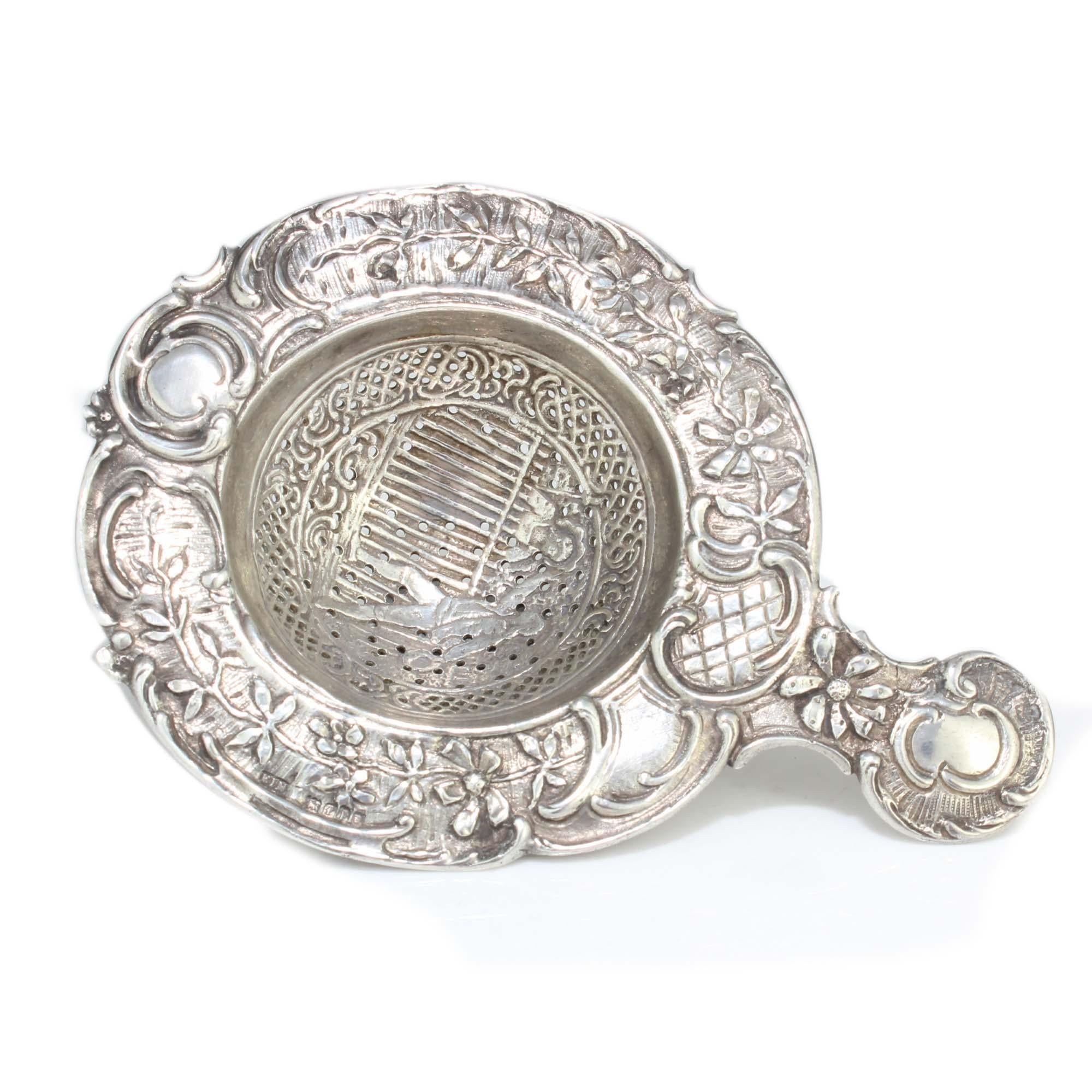 Antique silver tea strainer decorated with lady and gentleman, scrolls and flower garlands.
 
 Maker: Friedrich Reusswig 
 Made in Germany, 20th Century 
 Import marks: Elly Isaac Miller, England, London, 1901
 Fully hallmarked.

 Dimensions