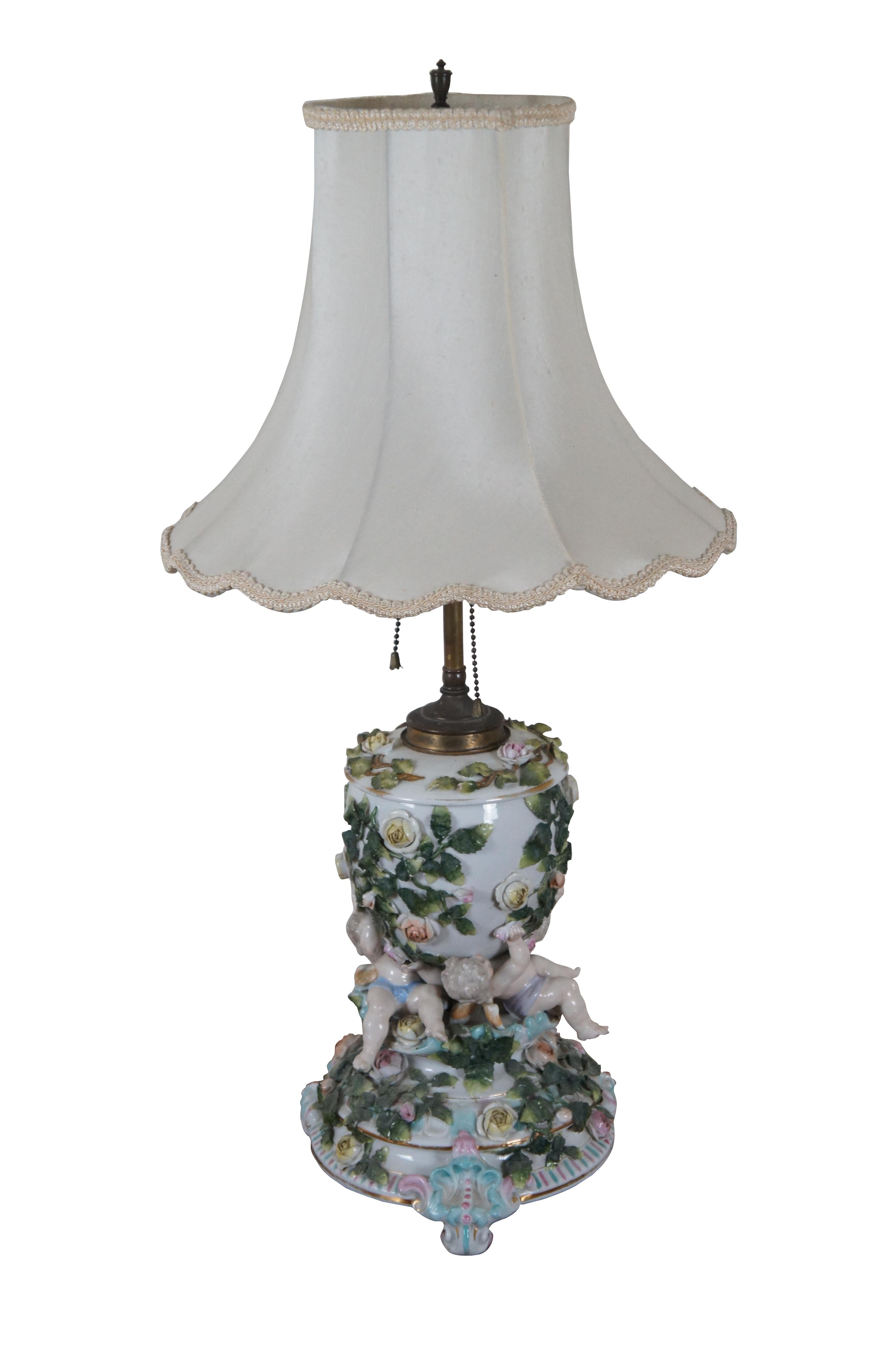 Antique German Sitzendorf Dresden converted oil lamp mantel trophy urn featuring a design of climbing roses and reclining cherubs. Marked on base with crossed lines, circa 1884 - 1902. Includes round white scalloped edge shade with braided