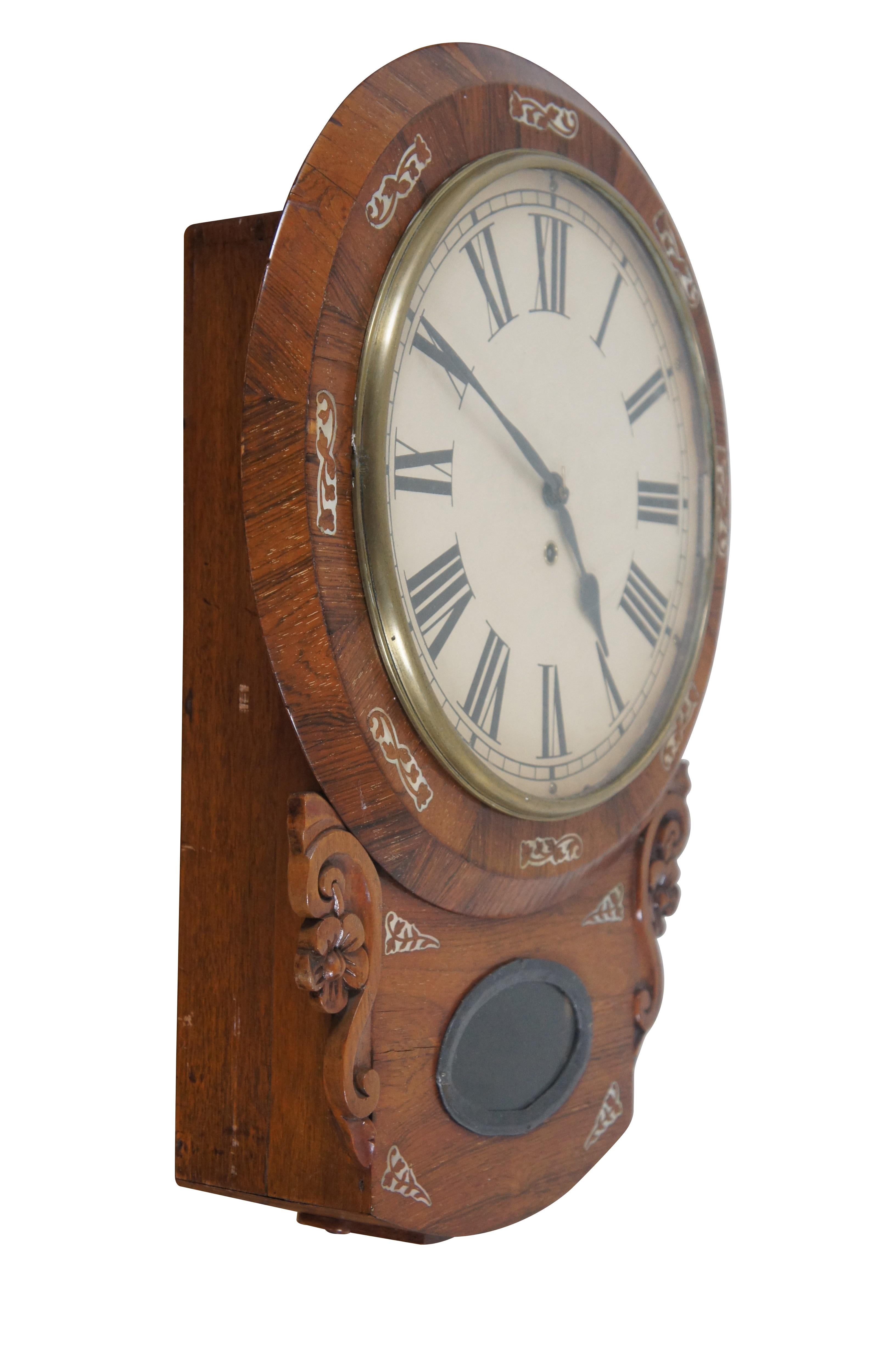 Antique short drop wall clock featuring a round beveled frame accented with inlaid mother of pearl cut-out leaf designs. The drop features an oval peekaboo window framed in black metal, showing the brass pendulum inside. Flanked by carved scrolls