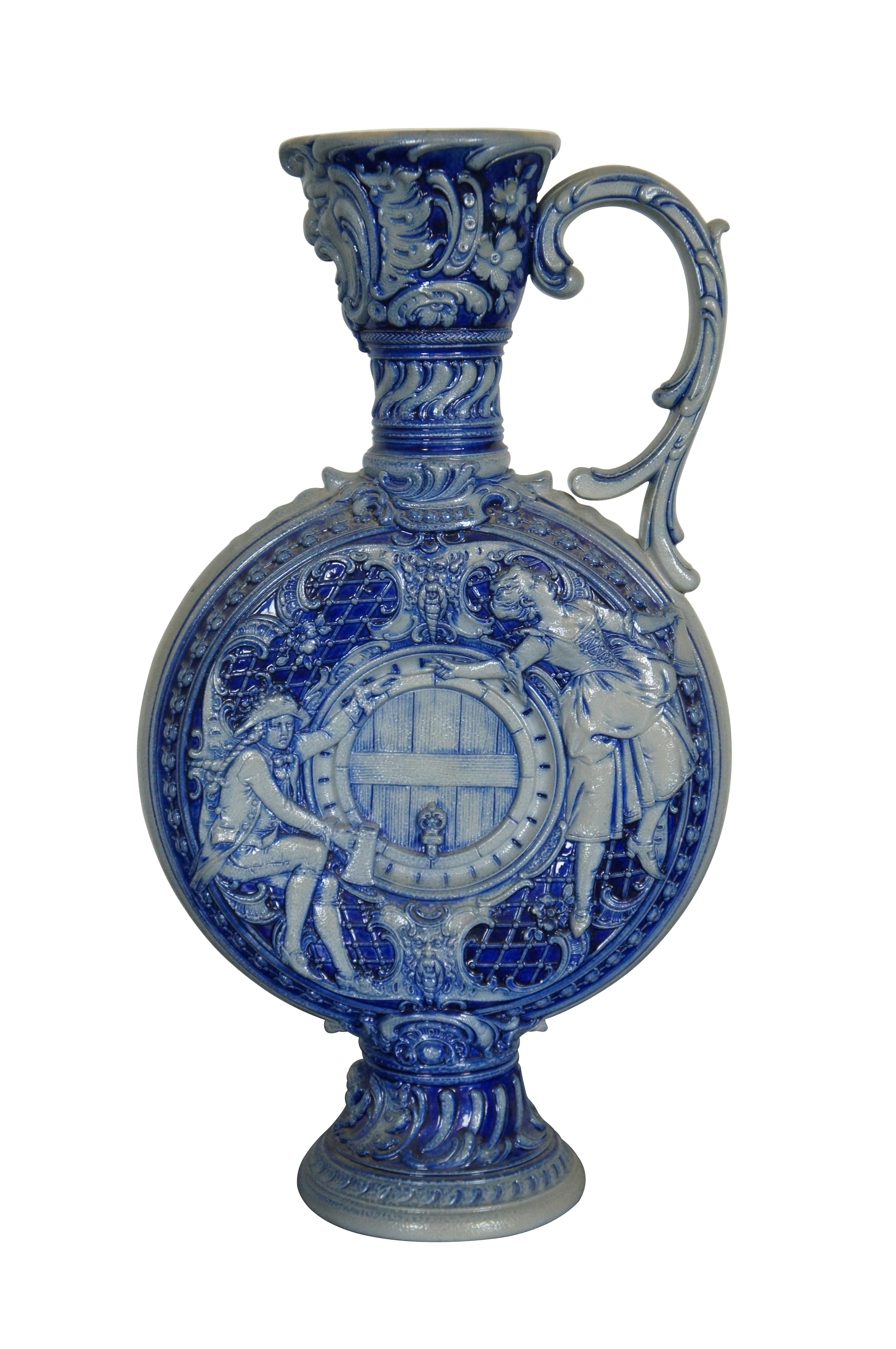 Ancienne carafe, aiguière ou pichet en poterie allemande en grès de Westerwald.  Réalisé en grès à glaçure salée bleu cobalt, il représente Dionysos / Bacchus, des chérubins, un homme ivre buvant à côté d'un tonneau et une femme dansant avec sa